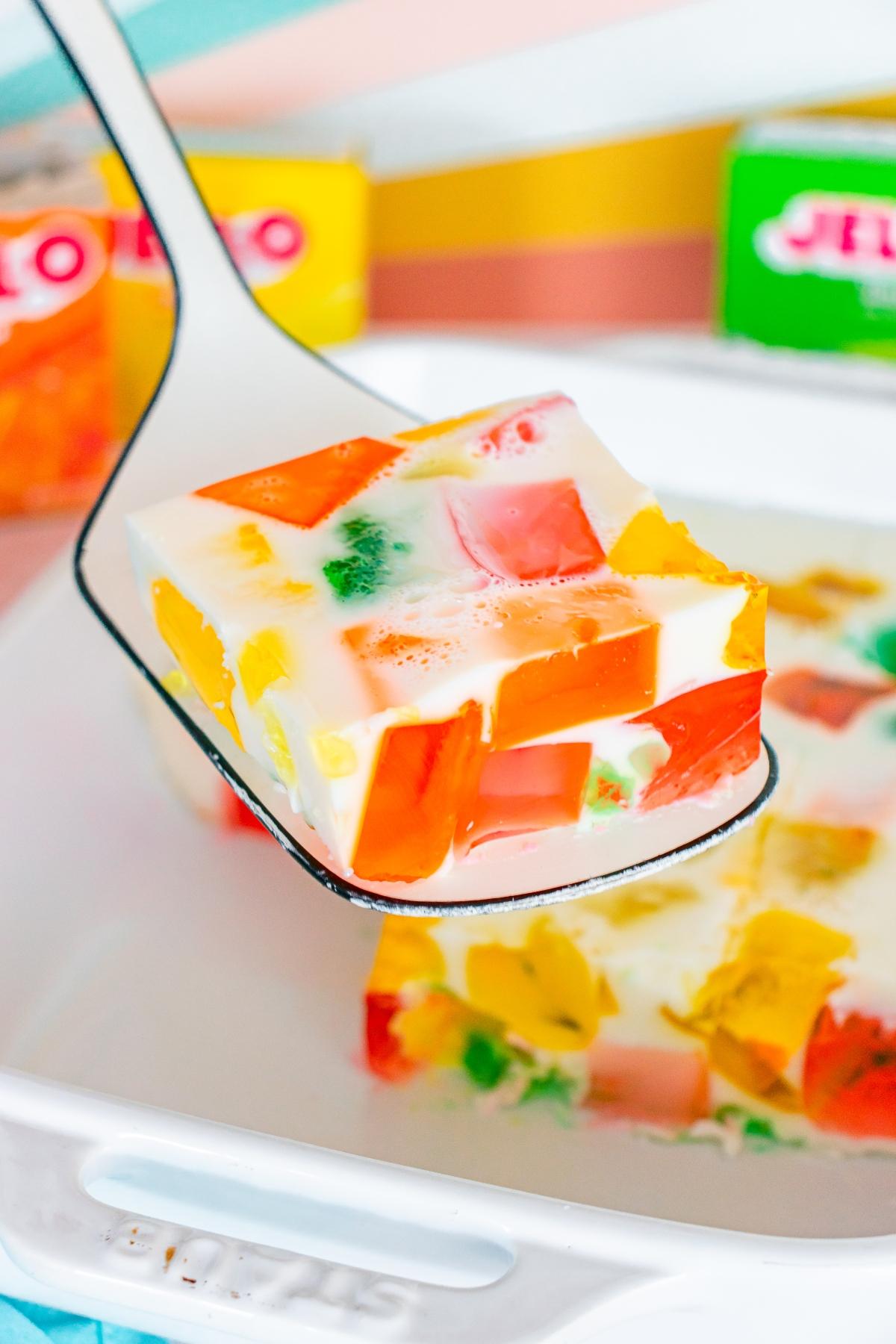 A spatula lifts a colorful Jello and cream dessert square from a white baking dish.