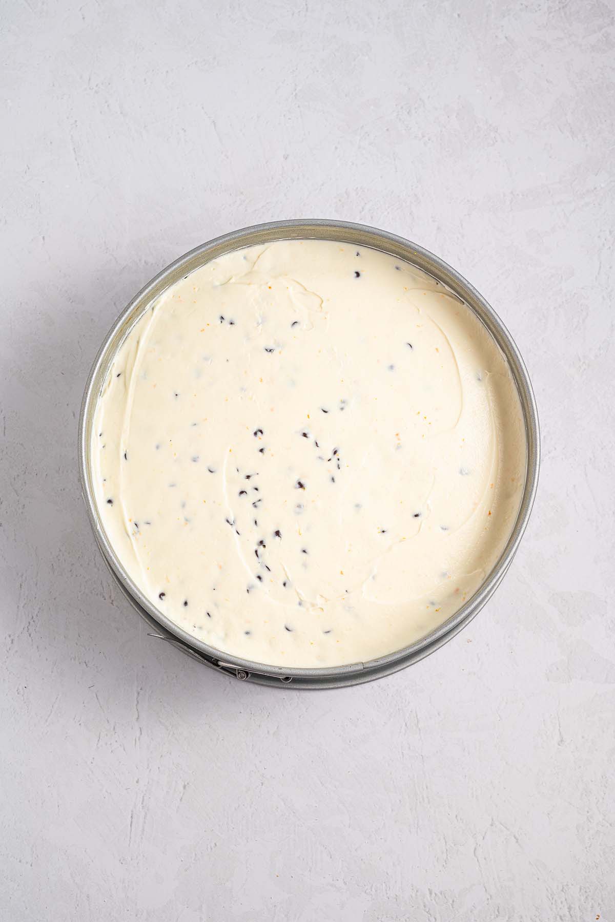 A round pan filled with creamy Cannoli Cheesecake batter and chocolate chips sits on a light textured surface.