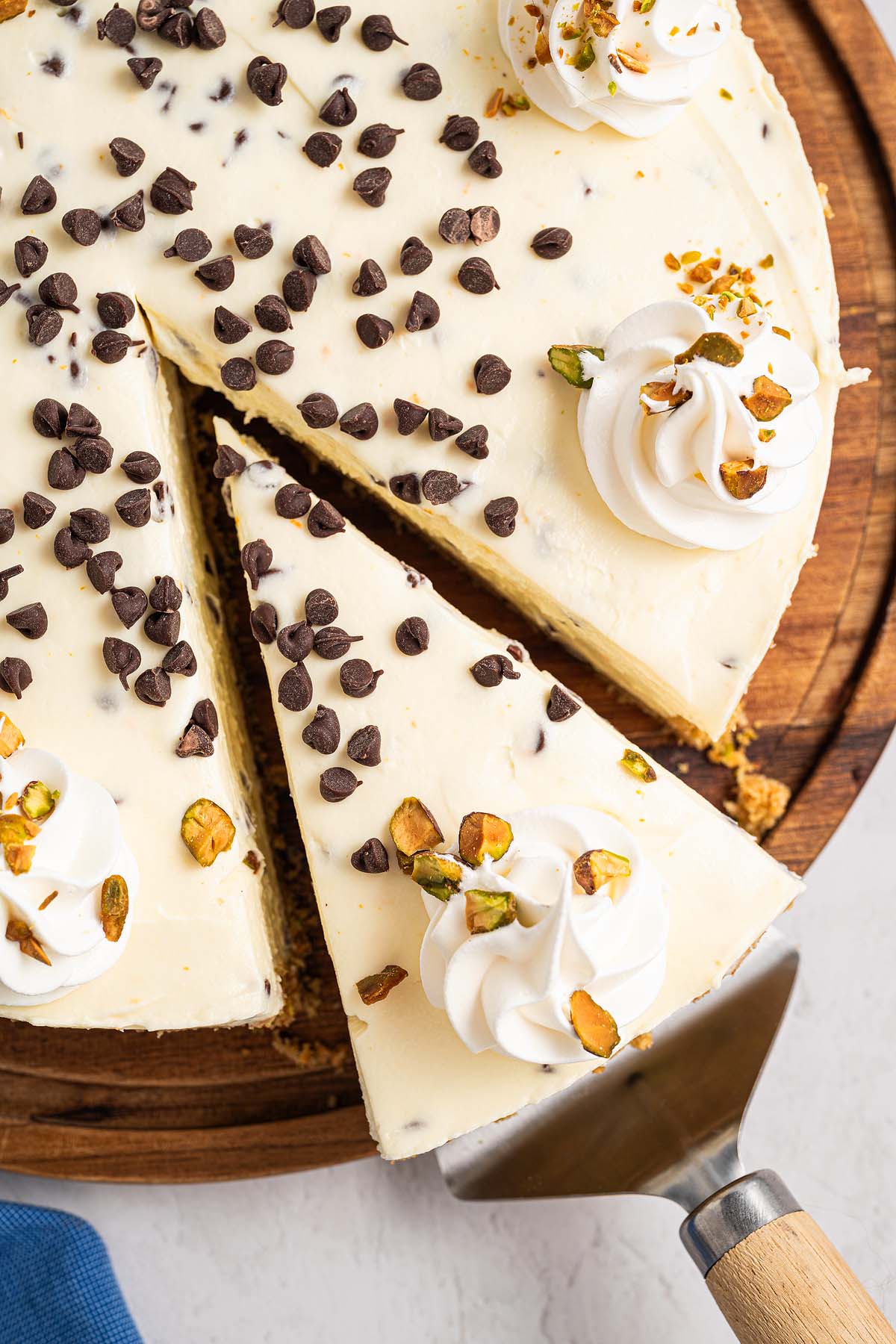 A Cannoli Cheesecake topped with whipped cream, chocolate chips, and chopped pistachios, with a slice being served.