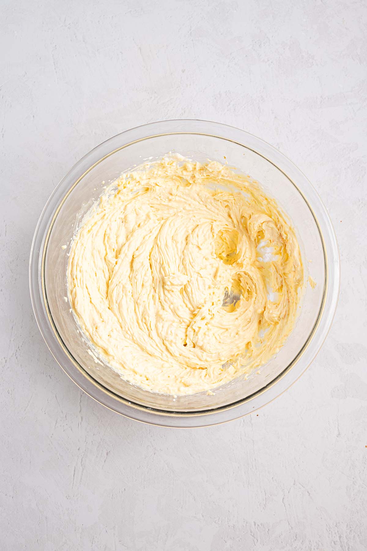 Glass bowl with creamy, whipped butter and sugar mixture for Cannoli Pie on a light textured surface.