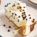 A slice of Cannoli Cheesecake with chocolate chips, nuts, and whipped cream on a plate with a fork.