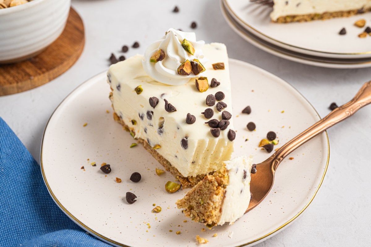 A slice of Cannoli Cheesecake with chocolate chips, chopped pistachios, and whipped cream on a plate with a fork.