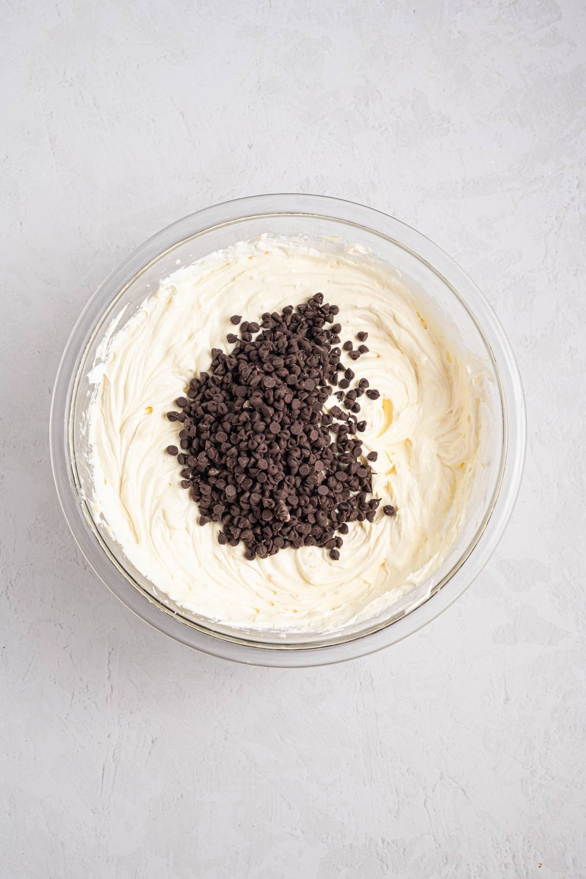 A glass bowl of whipped cream cheese with mini chocolate chips on top, ready to be mixed.