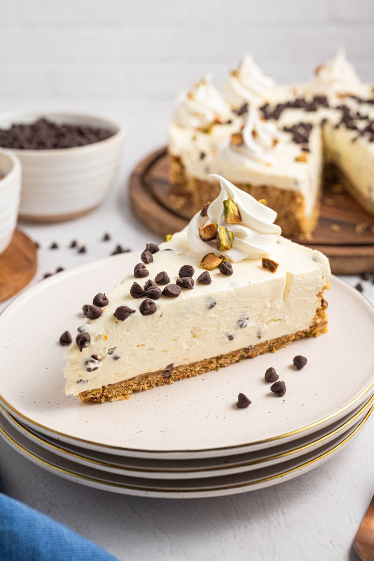 A slice of cannoli cheesecake topped with chocolate chips and pistachios on a plate, with the rest of the cake behind.