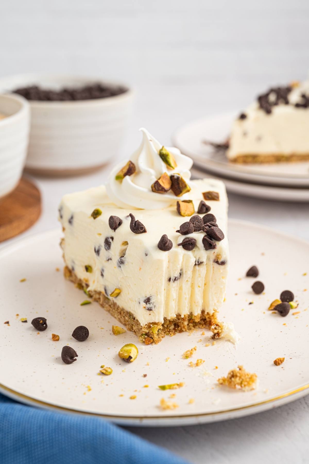A slice of cannoli cheesecake with chocolate chips and chopped pistachios, topped with whipped cream, on a white plate.