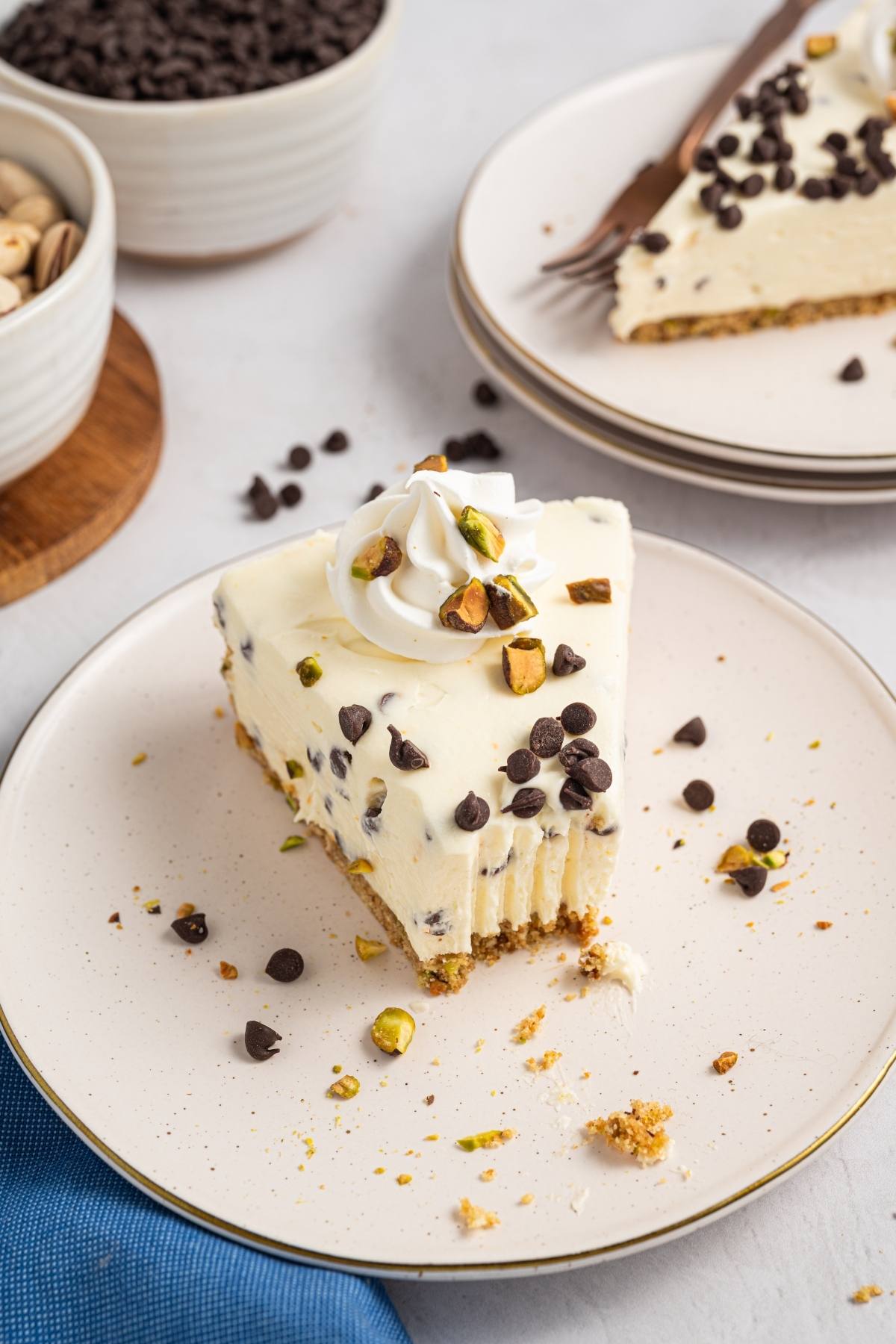 A slice of cannoli cheesecake topped with whipped cream, chocolate chips, and pistachios on a plate.