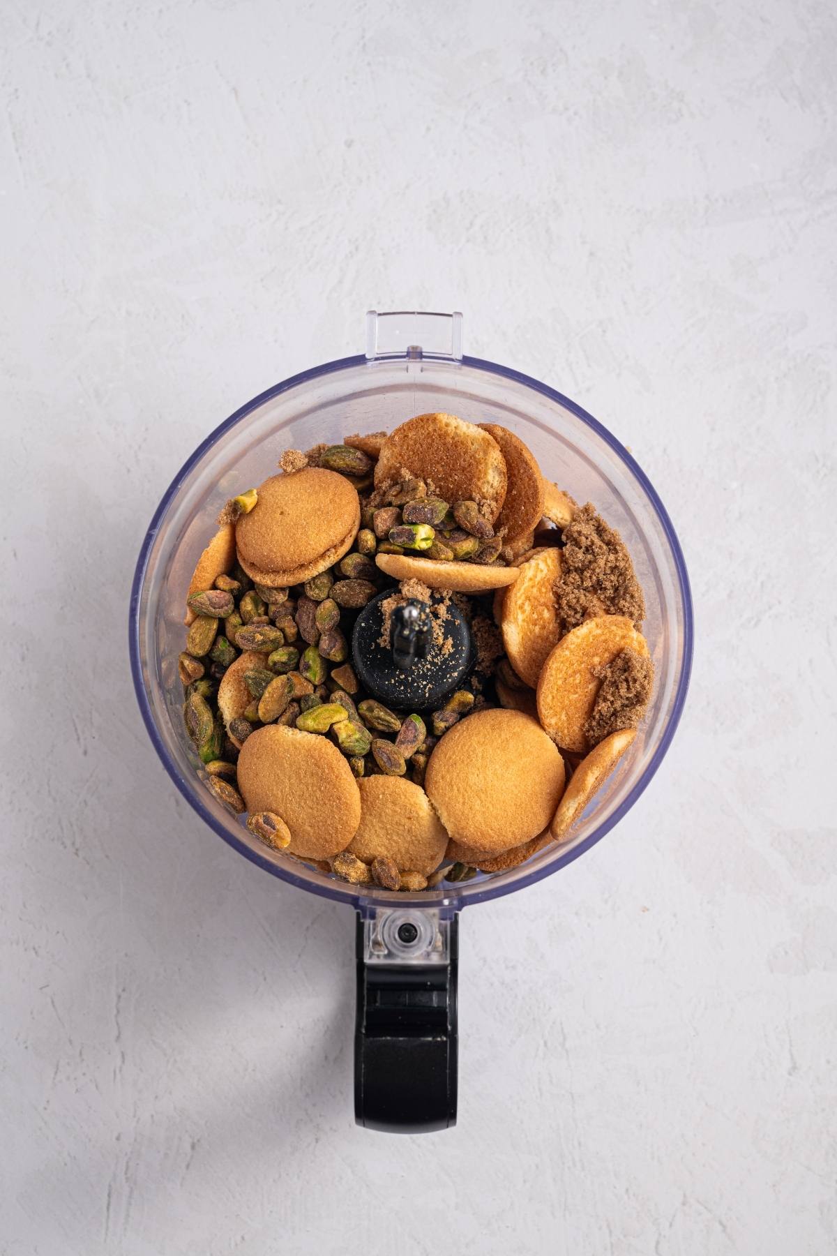 A food processor bowl filled with cookies, pistachios, and brown sugar on a light surface.