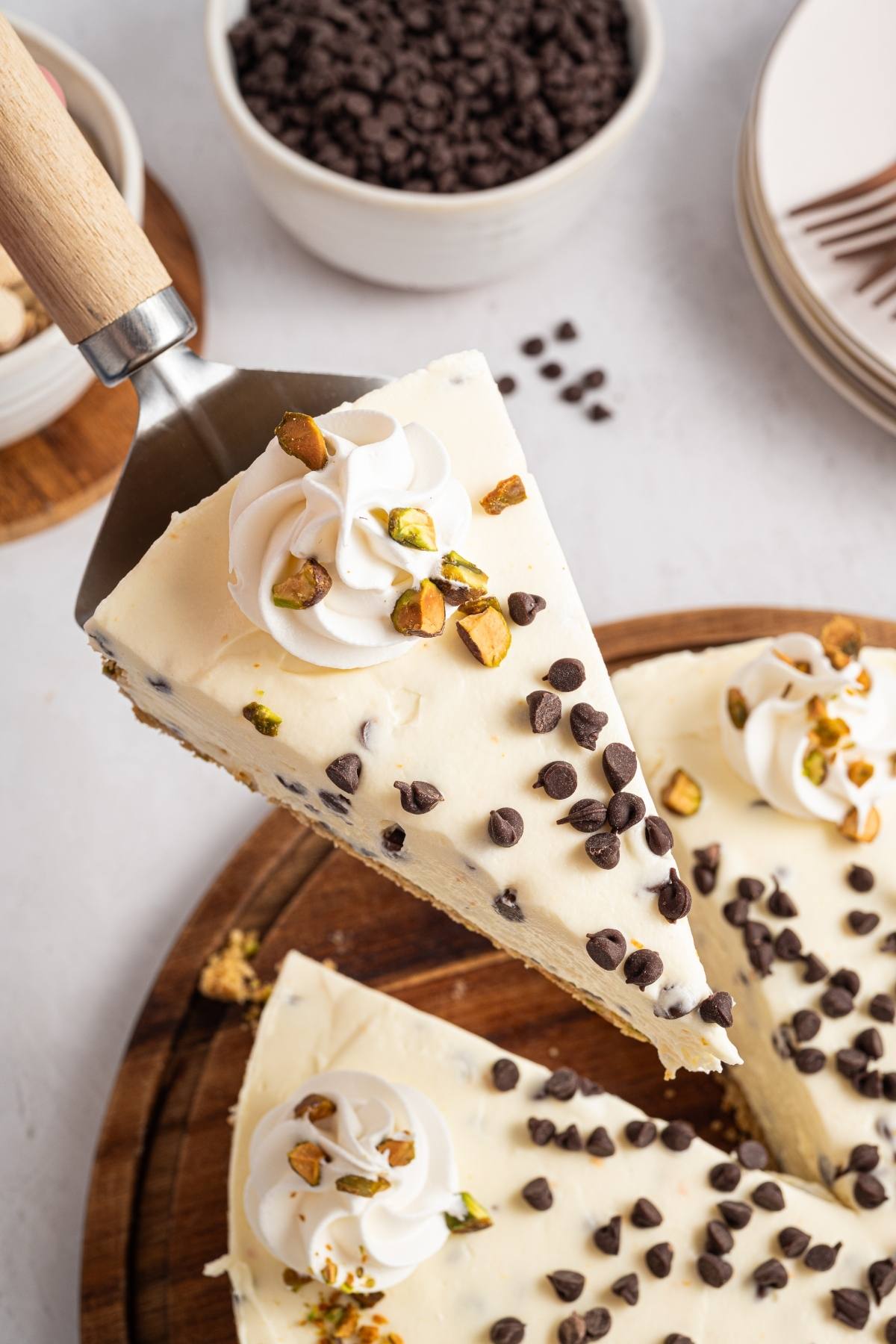 A slice of cannoli cheesecake topped with whipped cream, chocolate chips, and chopped pistachios on a cake server.
