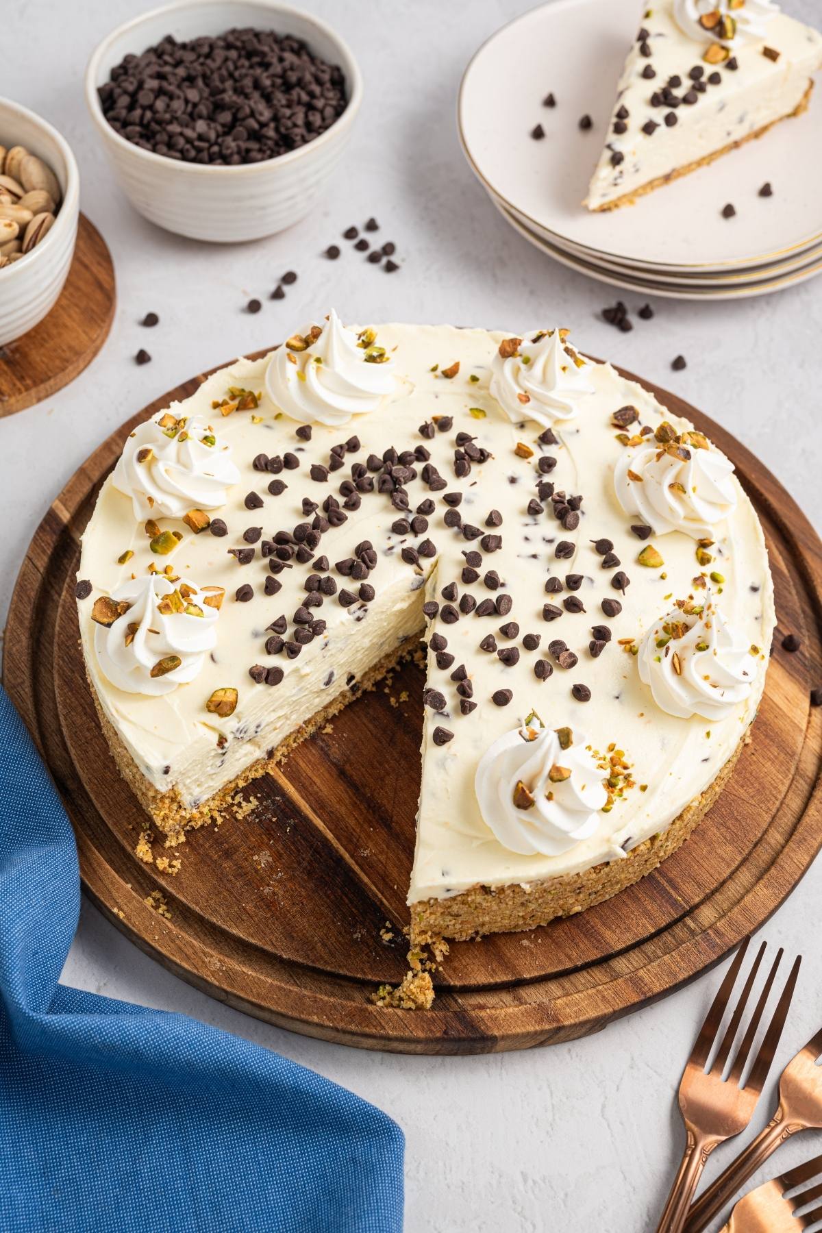 A cannoli cheesecake topped with whipped cream, chocolate chips, and pistachios, with one slice removed.