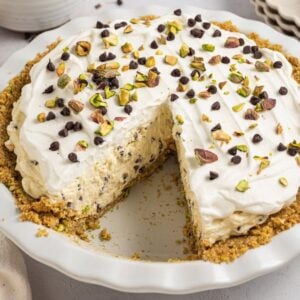 A Cannoli Pie with a slice missing, topped with whipped cream, chopped pistachios, and chocolate chips.
