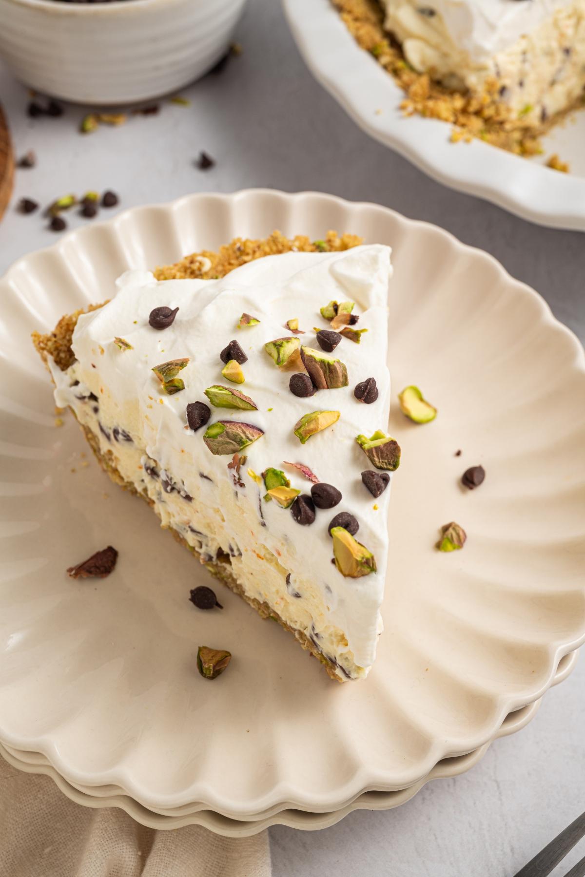 A slice of creamy pie topped with whipped cream, chocolate chips, and chopped pistachios on a scalloped plate.