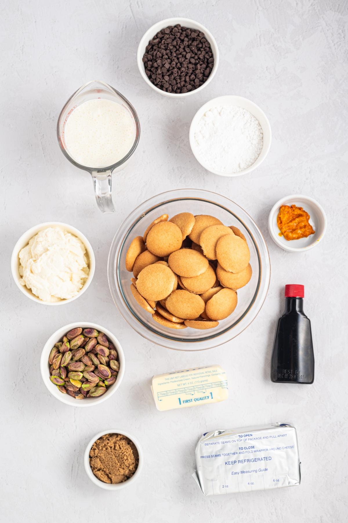 Bowl of vanilla wafers surrounded by small bowls of chocolate chips, nuts, cream, milk, spices, and vanilla.
