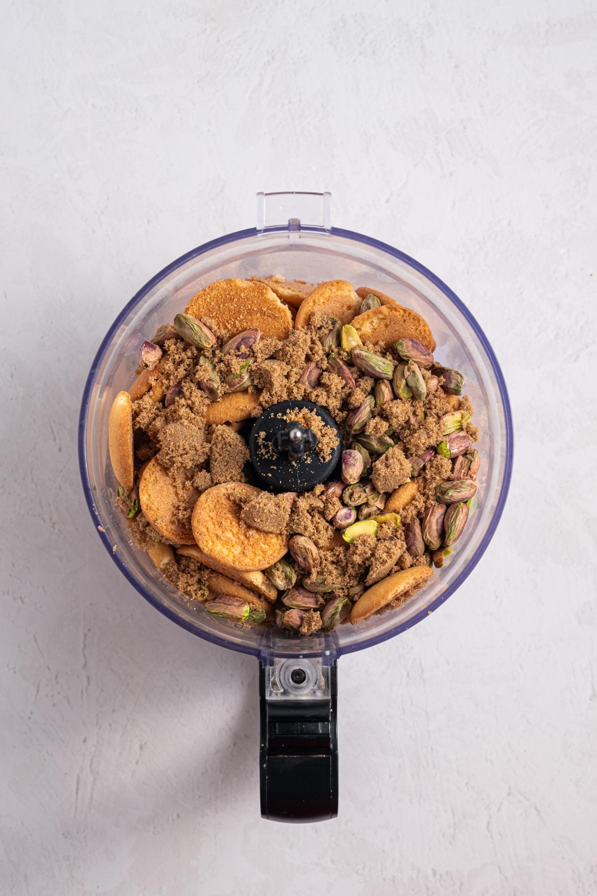 Overhead view of a food processor filled with pistachios, cookies, and brown sugar on a light surface.