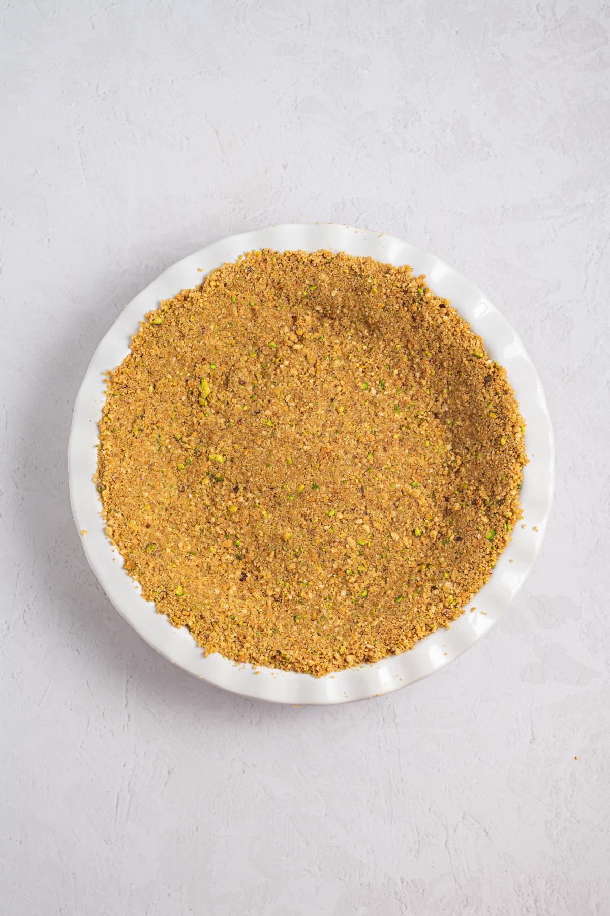 A pie dish filled with a pressed, crumbly, golden brown crust on a light textured surface.
