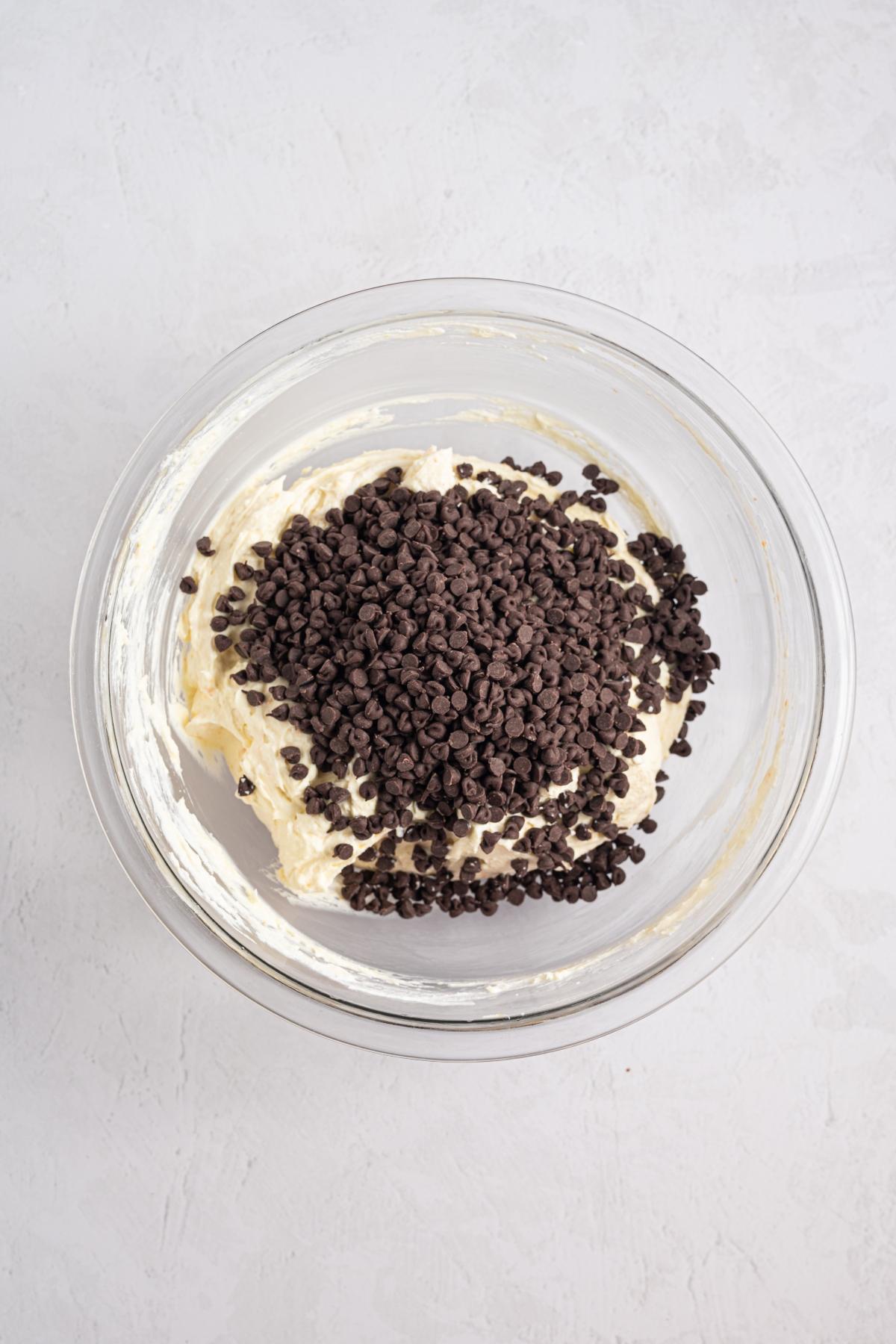 A glass bowl with cream cheese mixture topped with mini chocolate chips on a light surface.