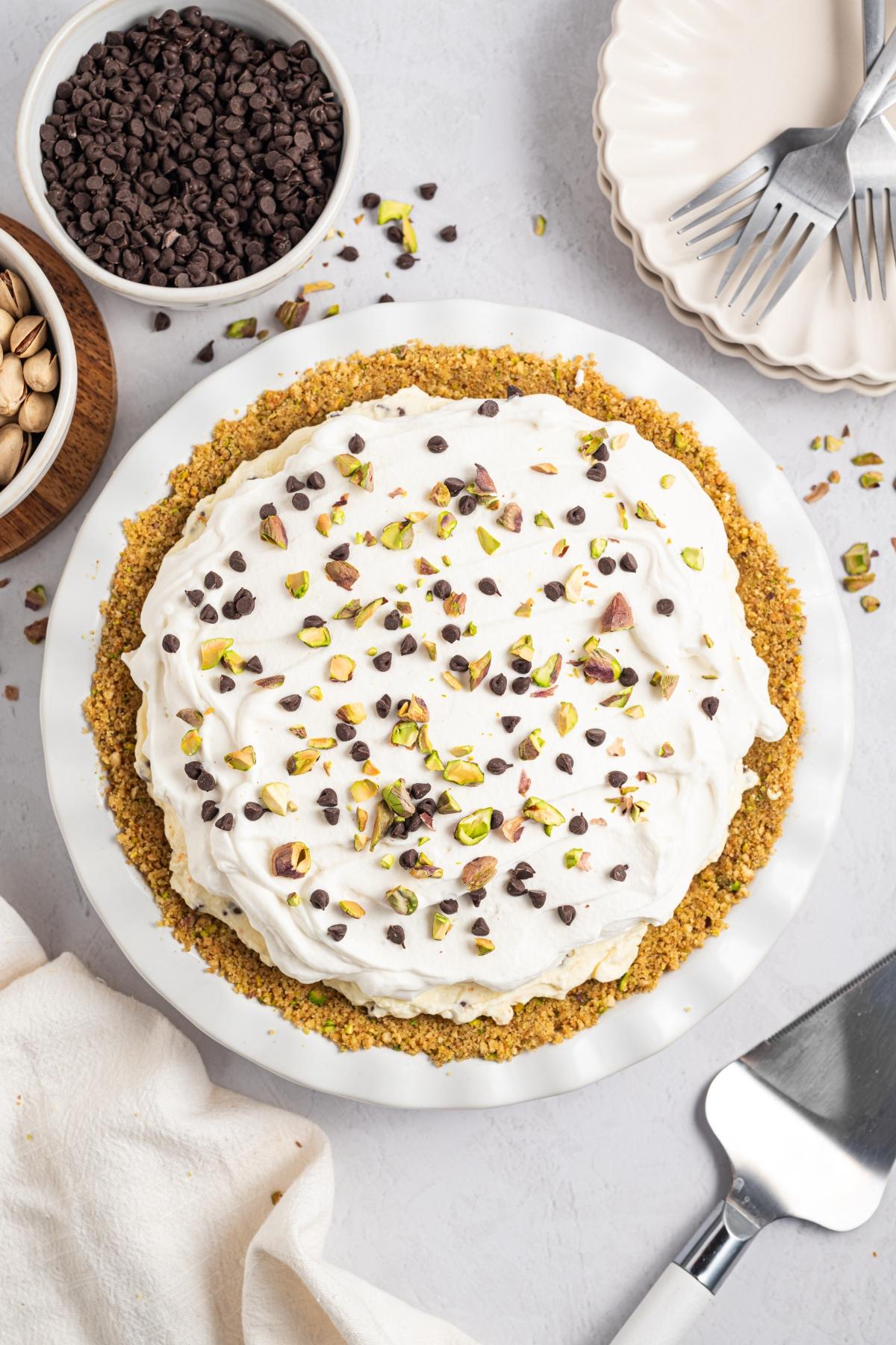 Pie topped with whipped cream, chopped pistachios, and chocolate chips, surrounded by bowls and utensils.