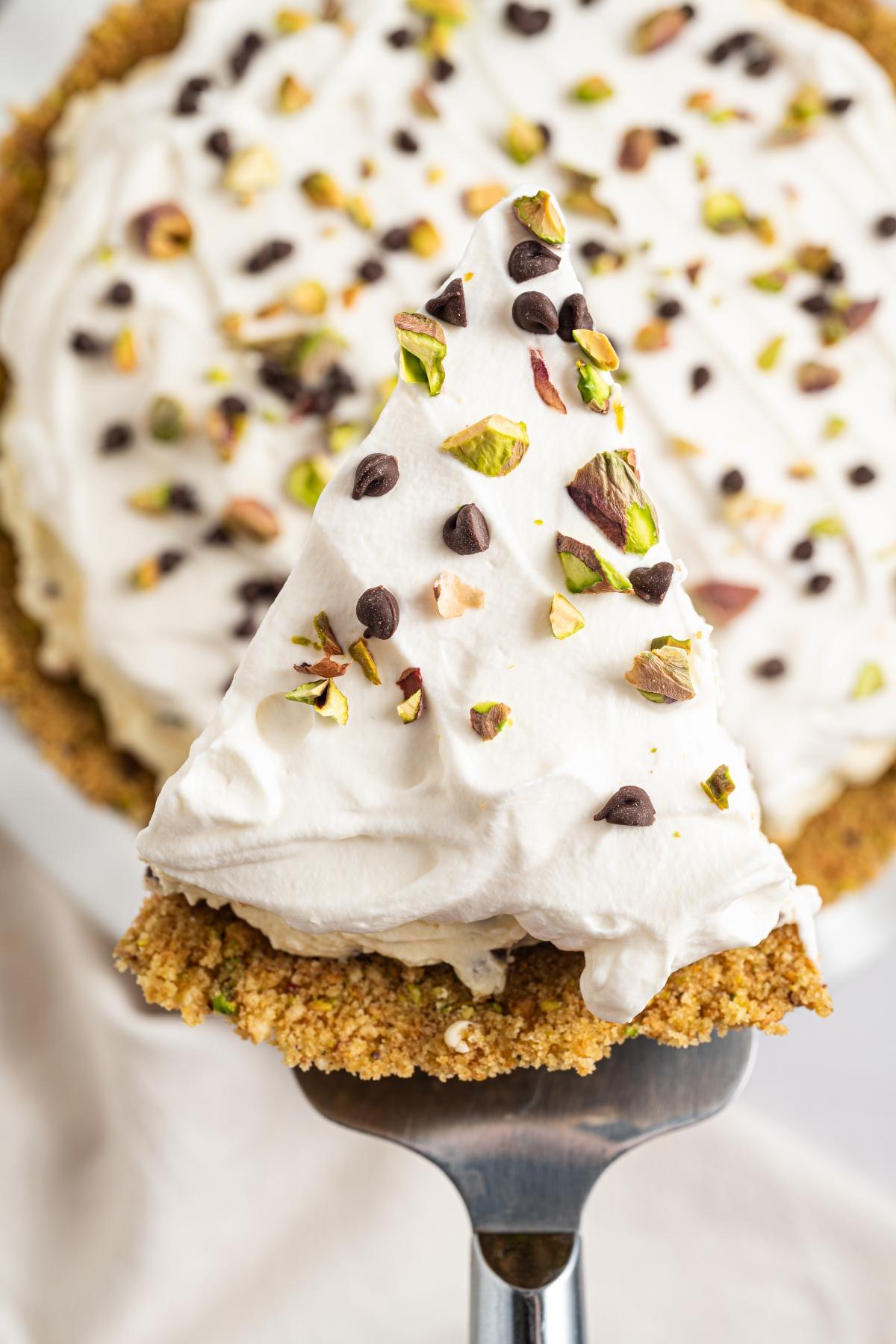 A slice of creamy pie with whipped topping, chocolate chips, and chopped pistachios on a pie server.