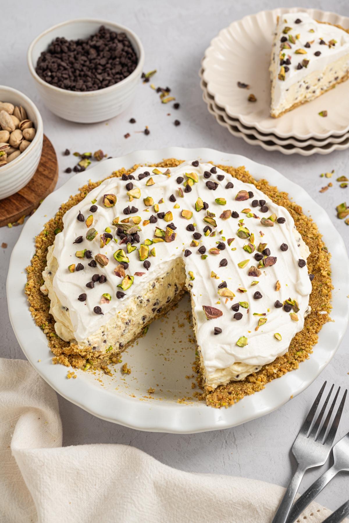 A creamy pie topped with whipped cream, chocolate chips, and pistachios, with a slice served on a plate nearby.