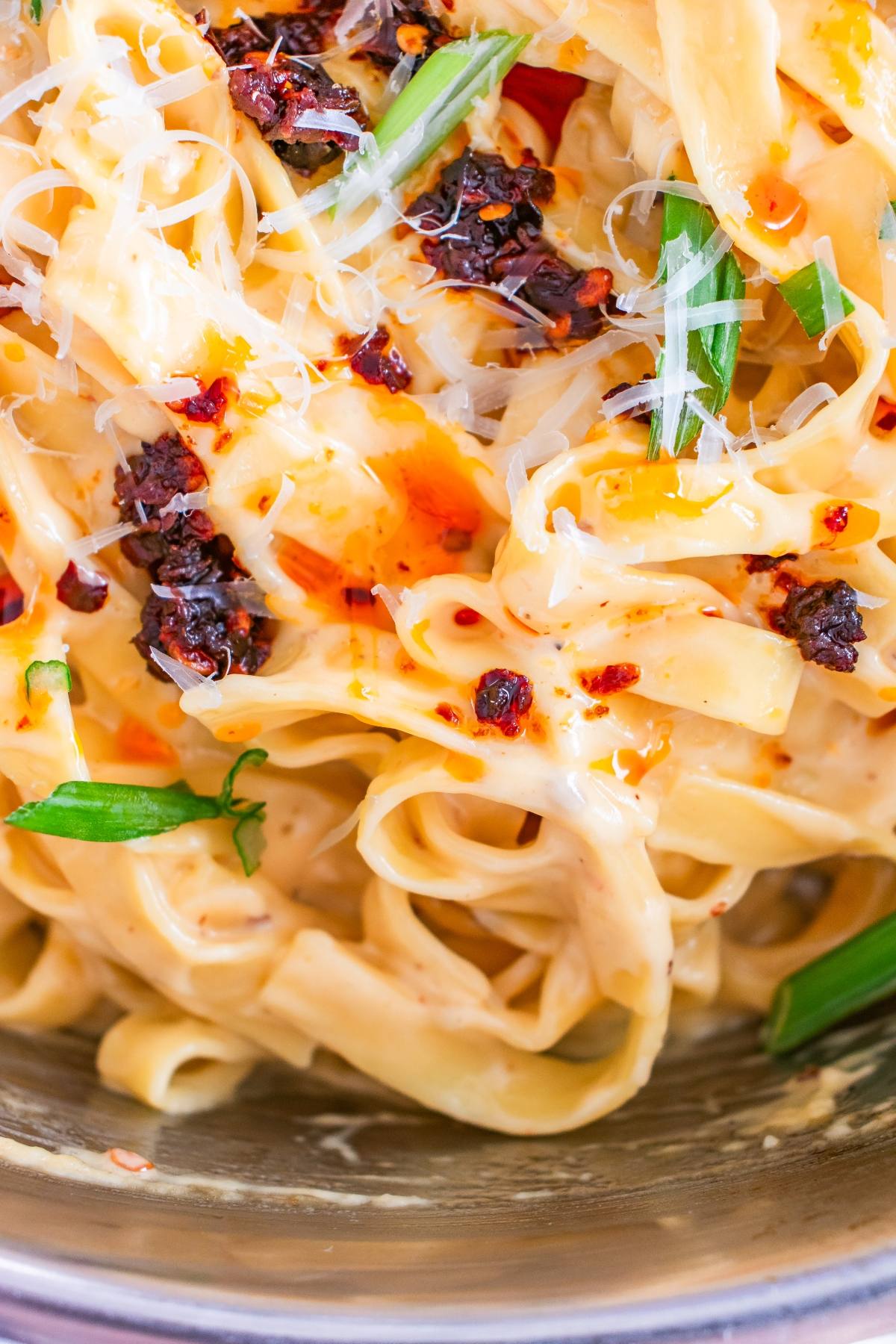 Close-up of creamy fettuccine pasta topped with chili oil, grated cheese, and fresh green herbs.