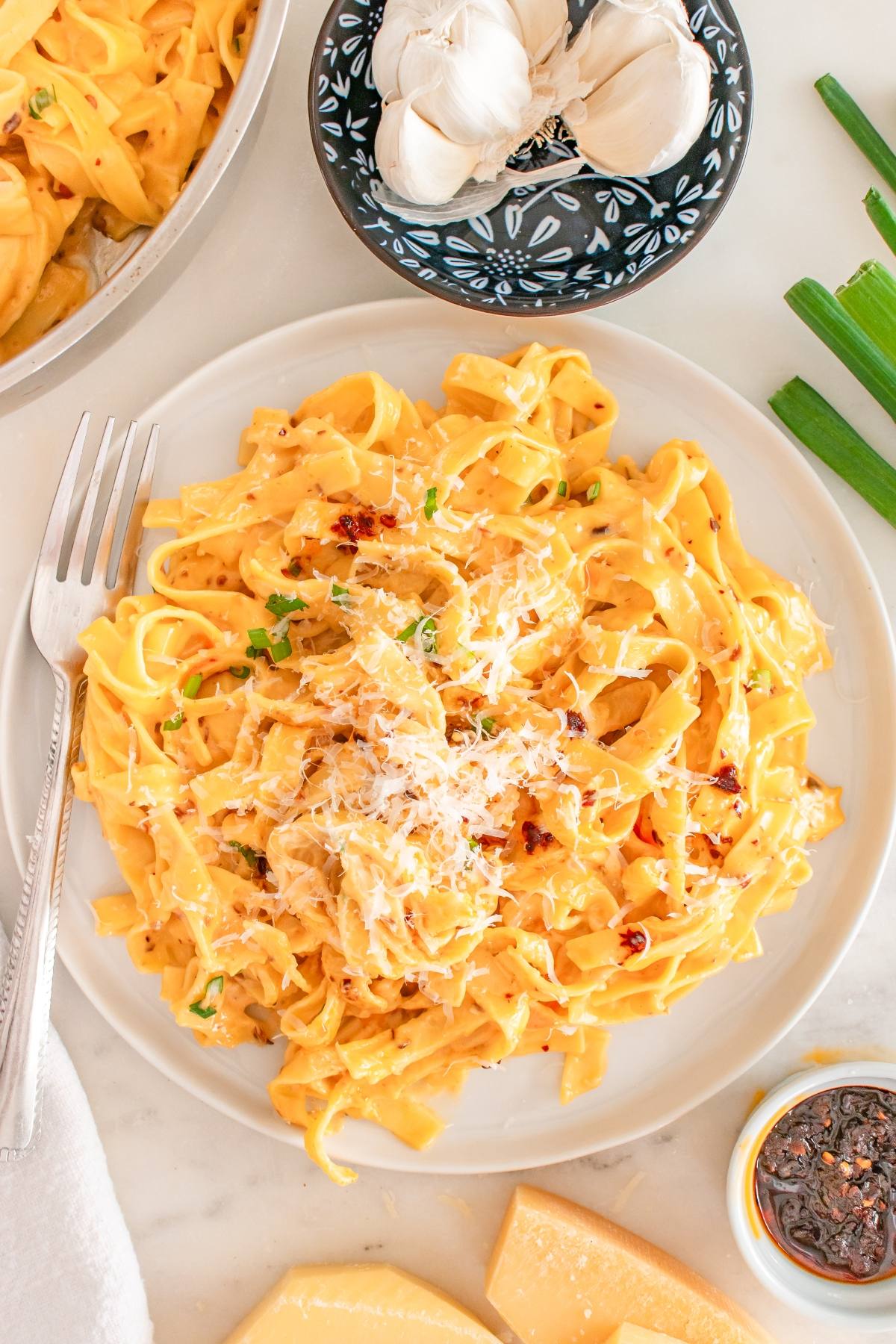 Plate of creamy fettuccine pasta topped with grated cheese and herbs, with garlic and sauce nearby.