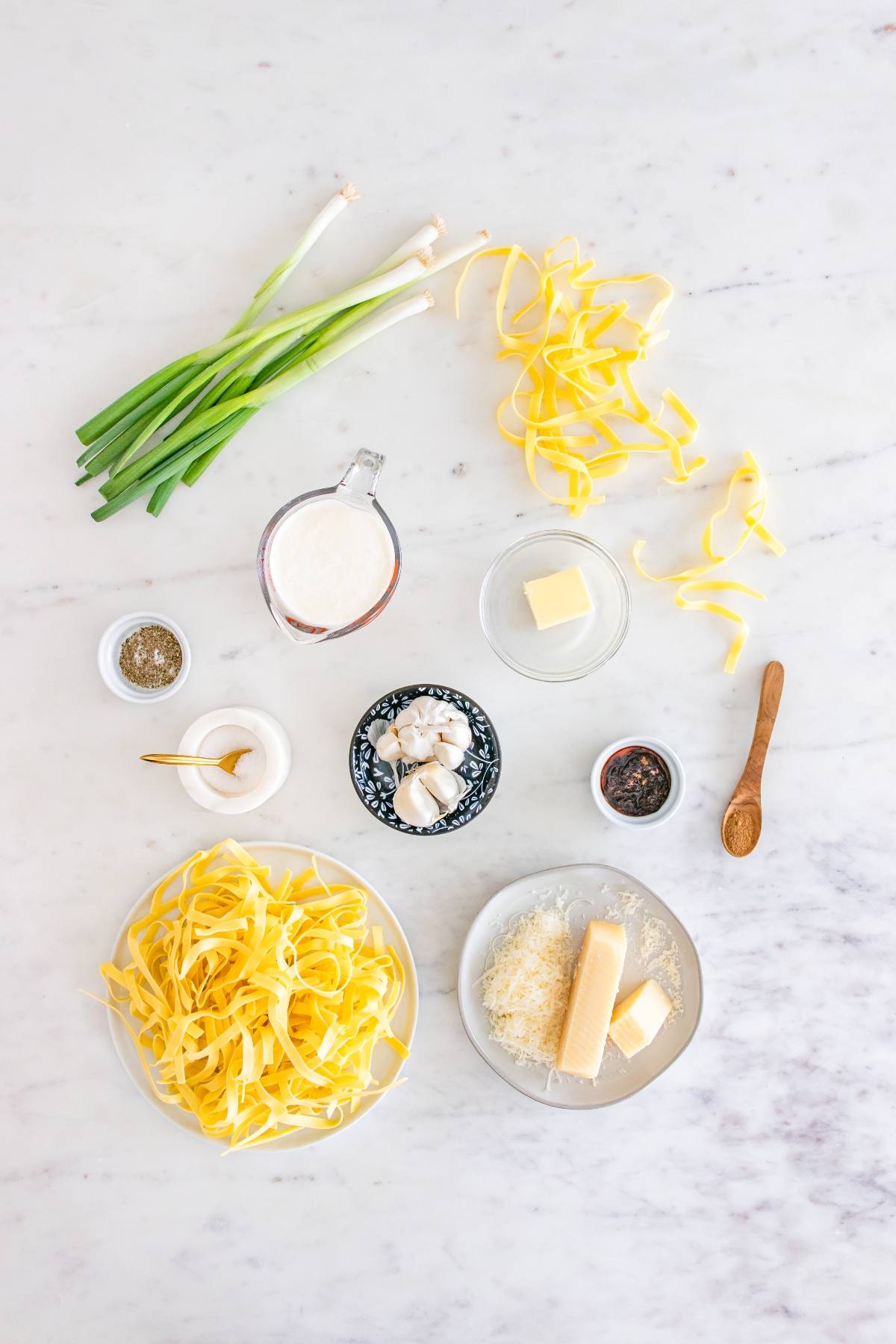 Ingredients for pasta Alfredo: tagliatelle, cream, butter, garlic, cheese, scallions, and seasonings on a table.