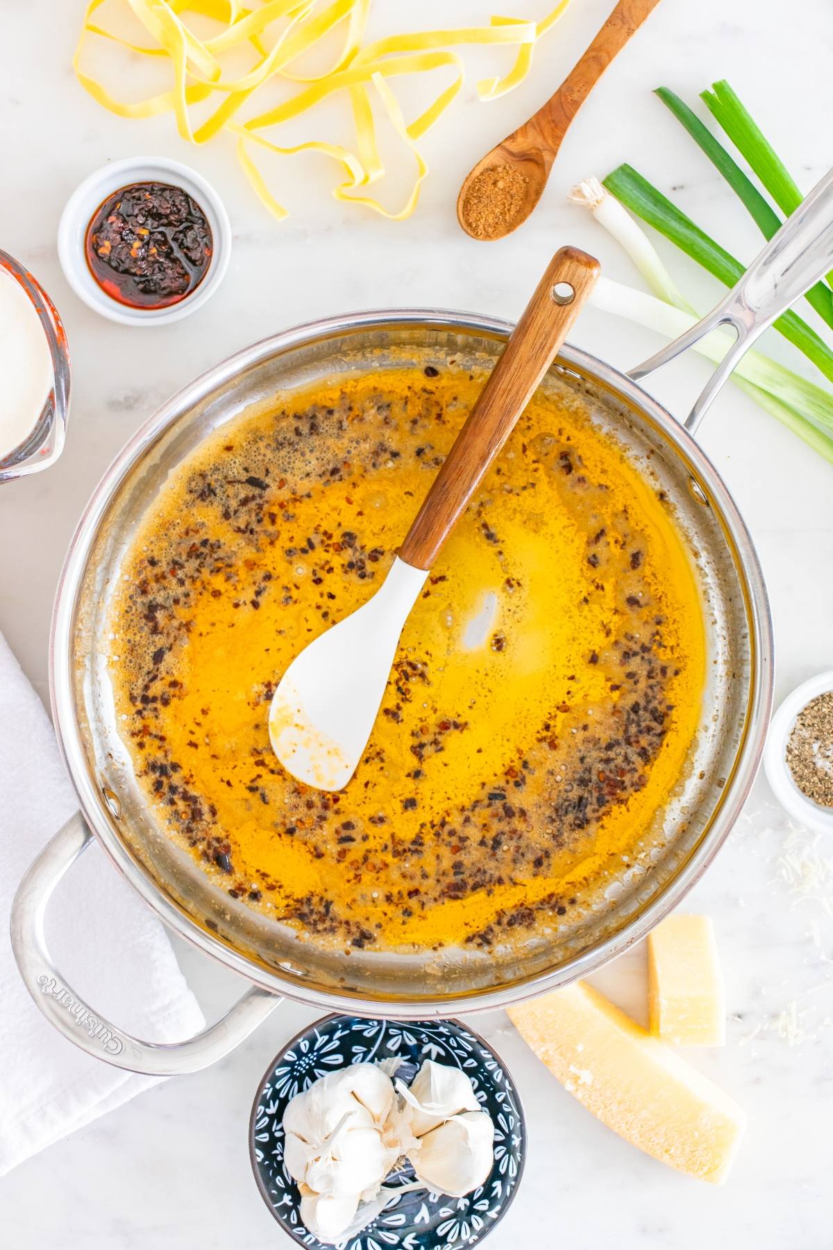 A pan of brown butter sauce with a spatula, surrounded by pasta, spices, cheese, and green onions.