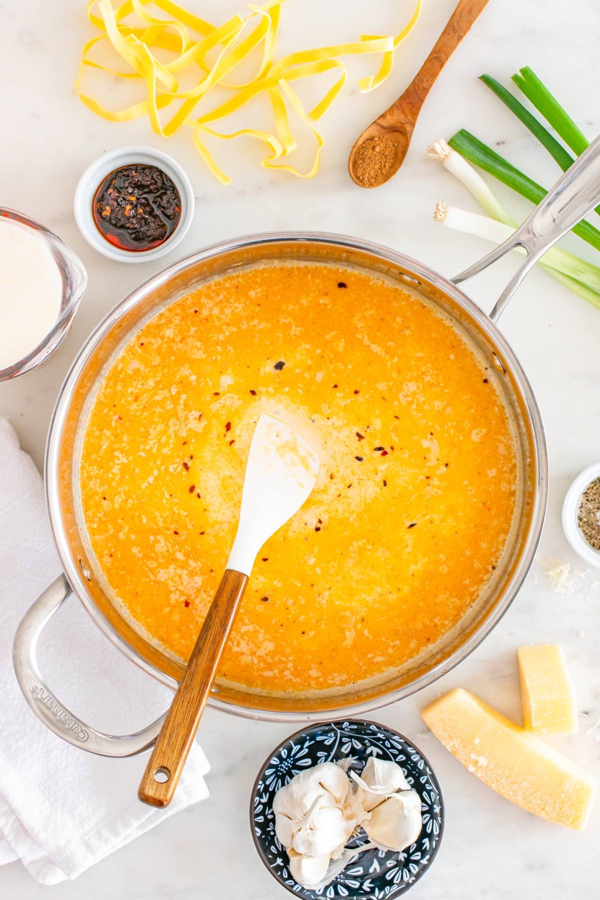 A pot of creamy orange sauce with a spatula, surrounded by pasta, cheese, garlic, and seasonings on a counter.