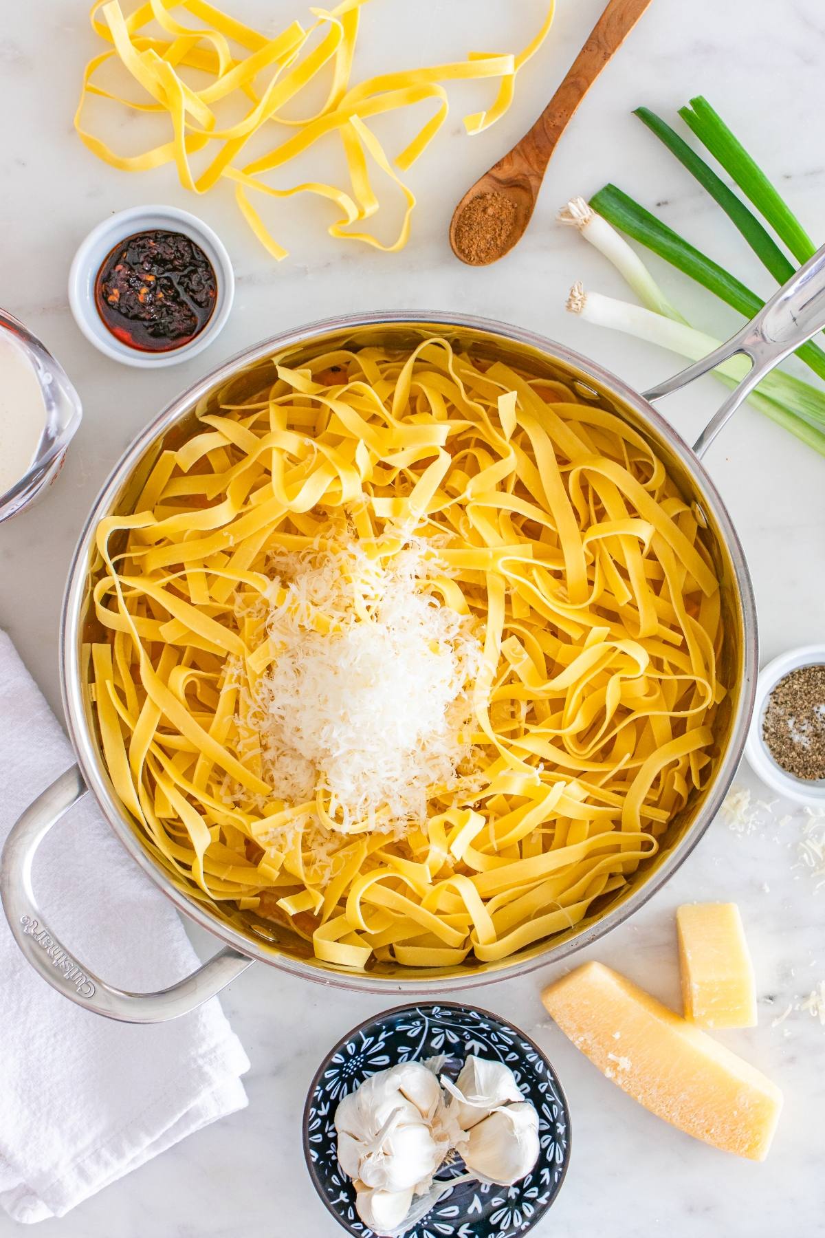 A pot of uncooked tagliatelle pasta with grated cheese, surrounded by garlic, cheese, spices, and green onions.