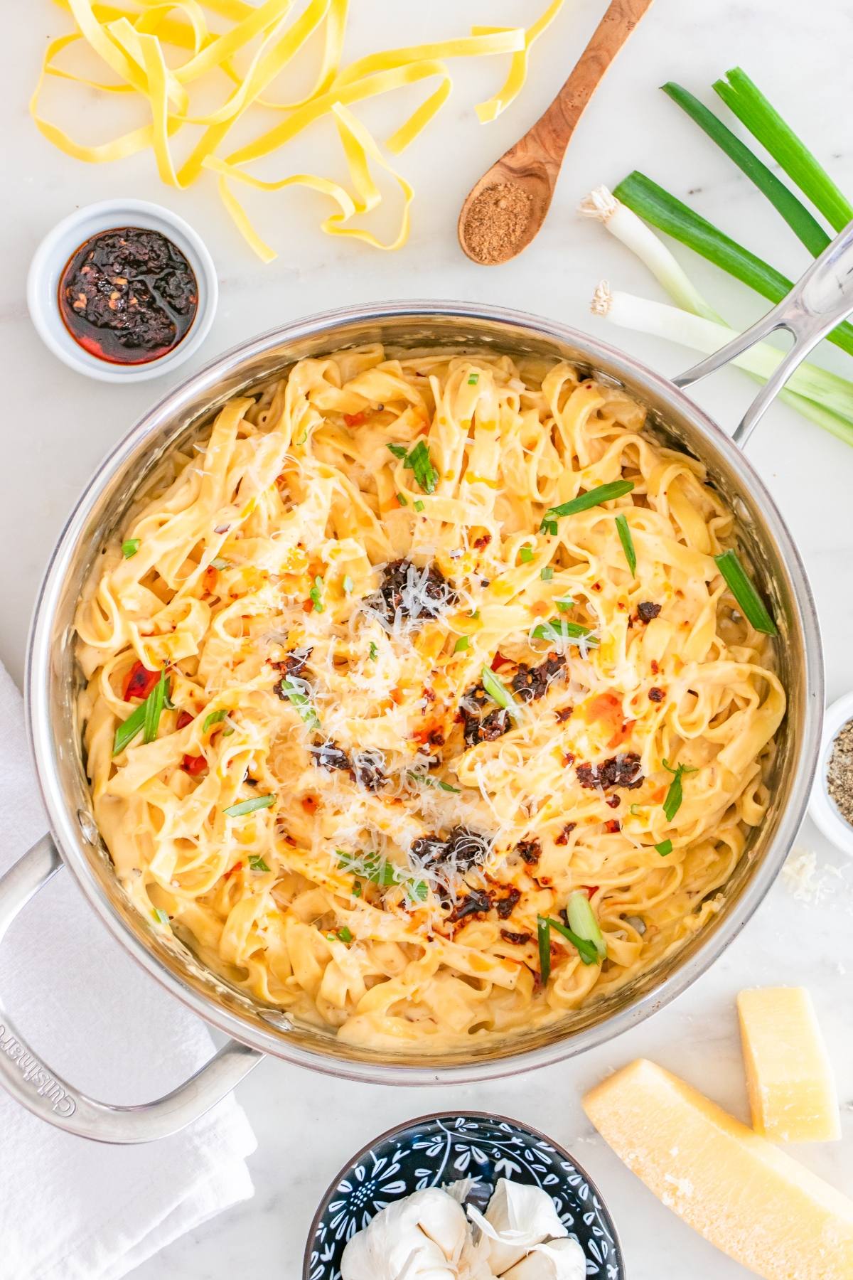Creamy pasta in a pan topped with chili oil, green onions, and cheese, surrounded by ingredients on a white surface.