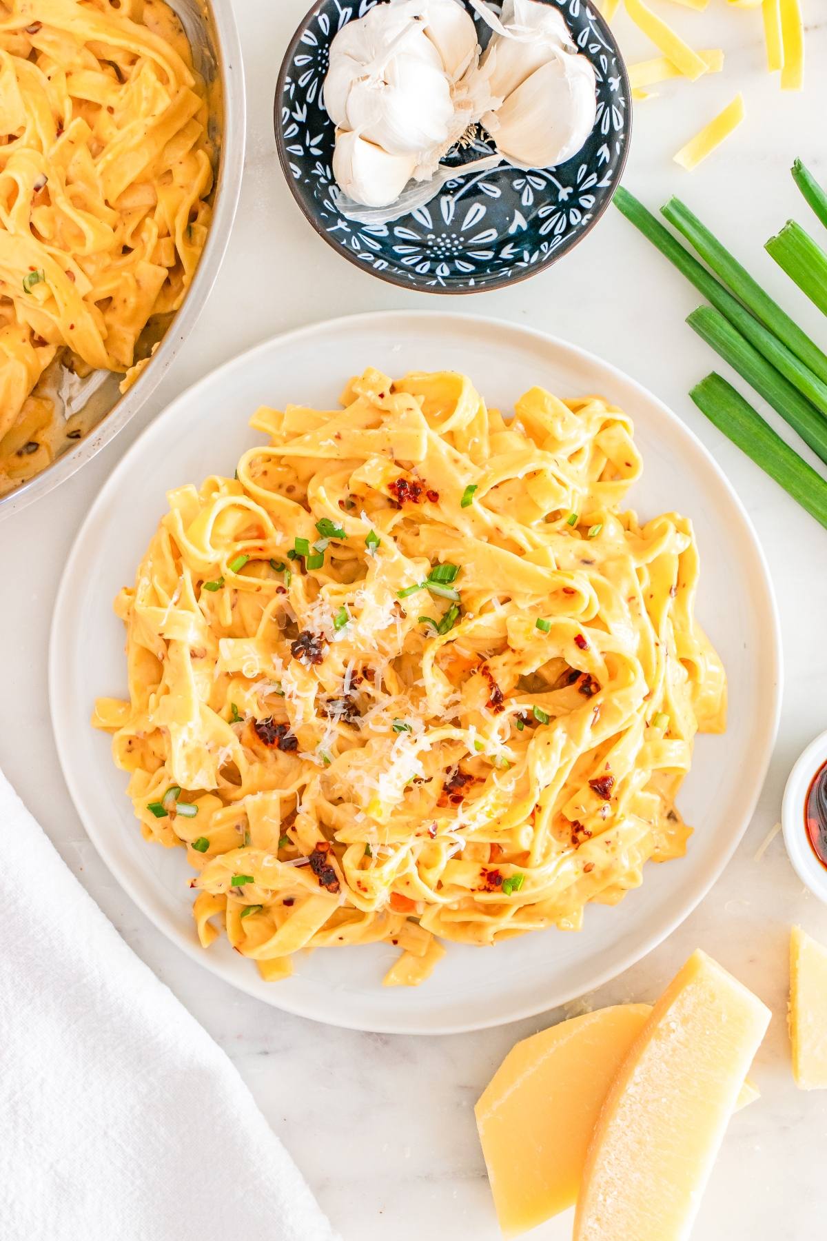 A plate of creamy fettuccine pasta topped with cheese and herbs, surrounded by garlic, cheese, and green onions.
