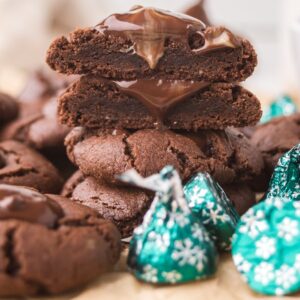Chocolate Mint Kiss Cookies with melted centers are stacked among green-wrapped chocolate candies on a table.