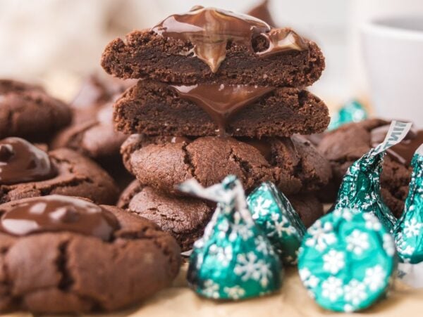 Chocolate Mint Kiss Cookies with melted centers are stacked among green-wrapped chocolate candies on a table.