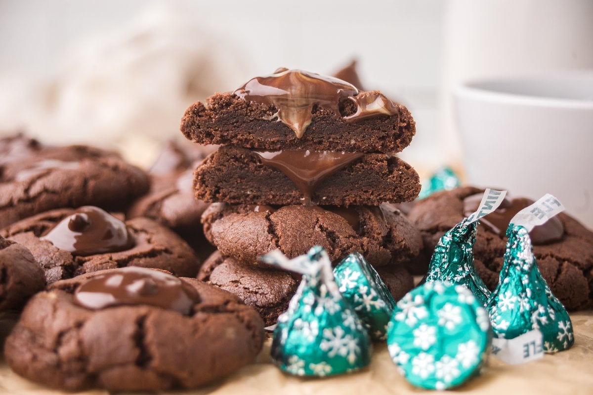 A stack of chocolate mint kiss cookies with melted chocolate and wrapped green-foil Chocolate Mint Kiss Cookies nearby.