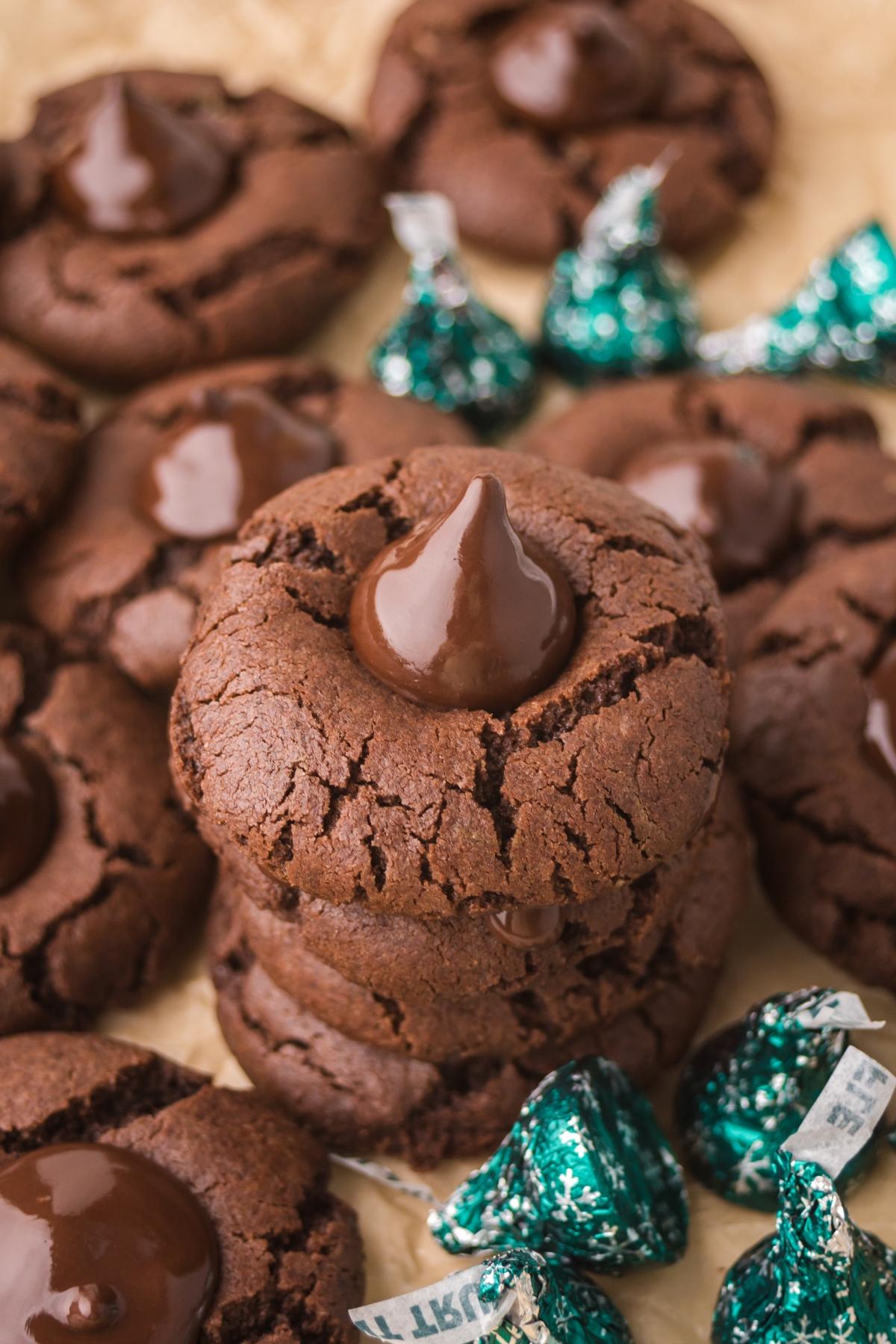 Chocolate mint kiss cookies with chocolate kisses on top, surrounded by wrapped chocolate candies on a brown surface.