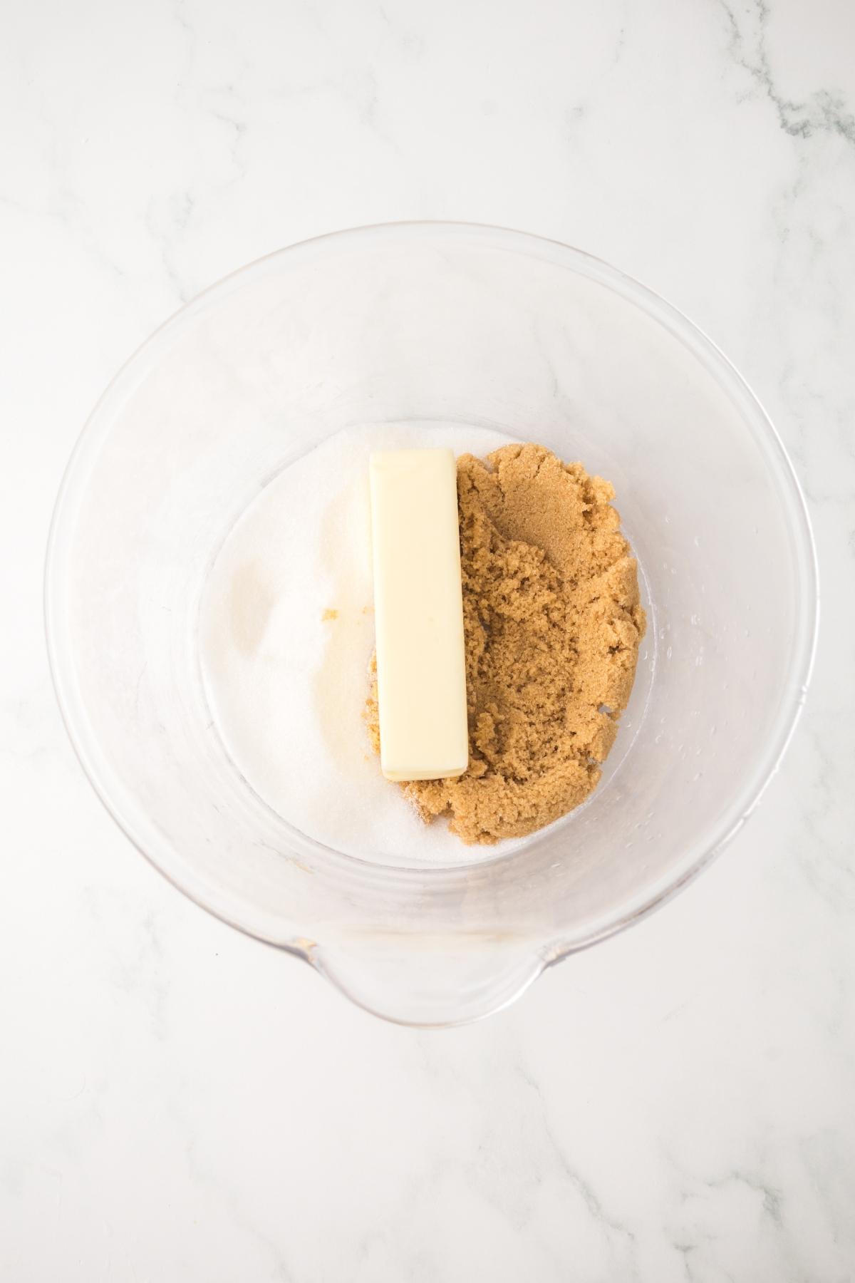 A glass bowl with a stick of butter, brown sugar, and white sugar on a white marble surface.