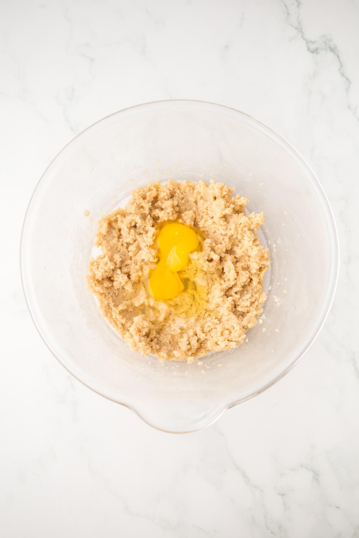 Clear bowl with creamed sugar and butter, plus two cracked eggs, on a white marble surface.