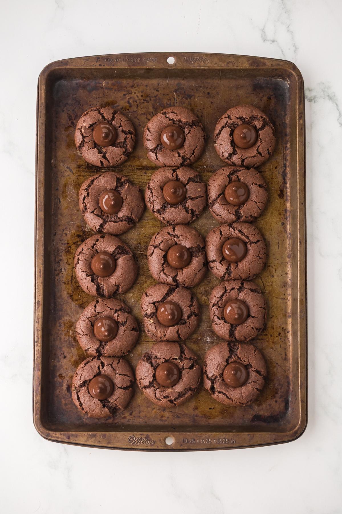 A baking sheet with fifteen chocolate mint cookies, each topped with a chocolate candy, on a white surface.