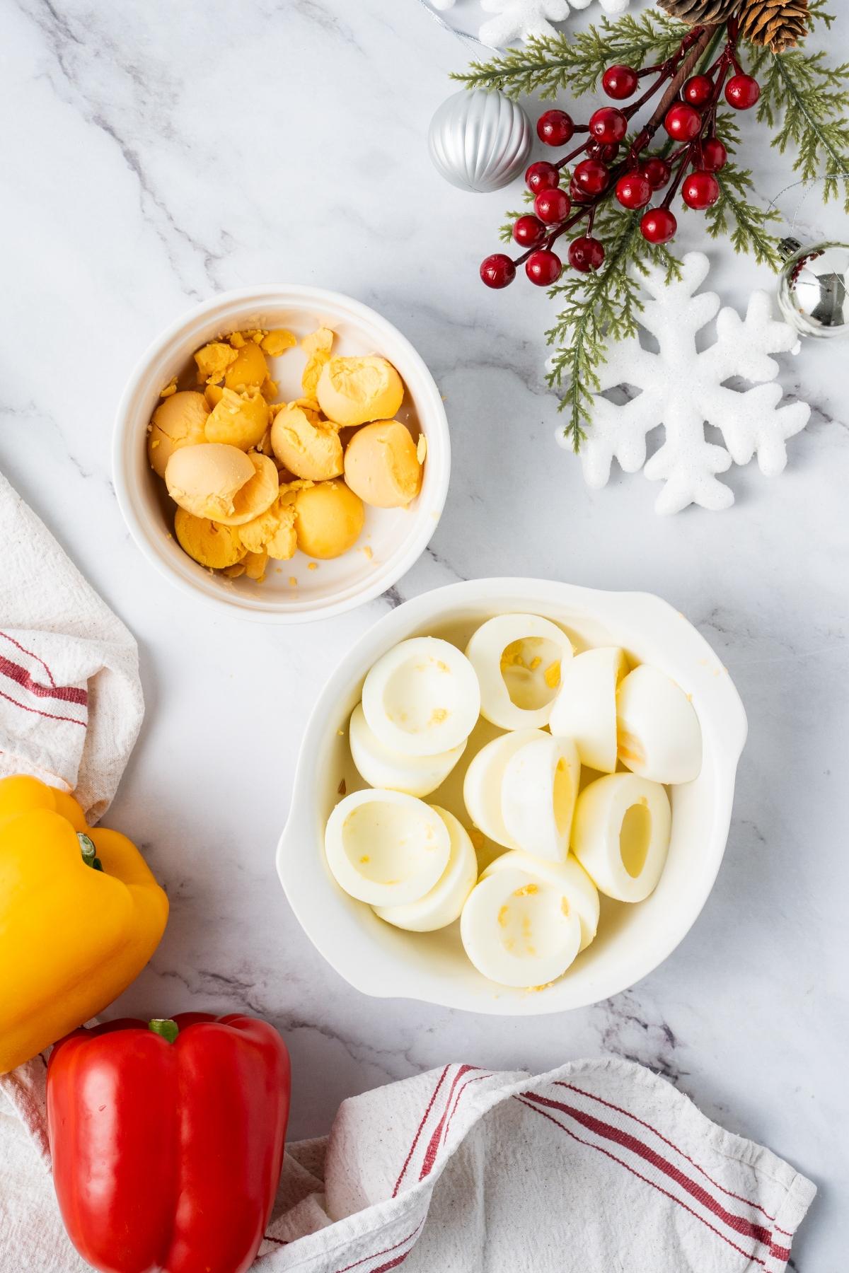Hollowed boiled egg whites and egg yolks in bowls on a marble surface with holiday decorations and bell peppers.