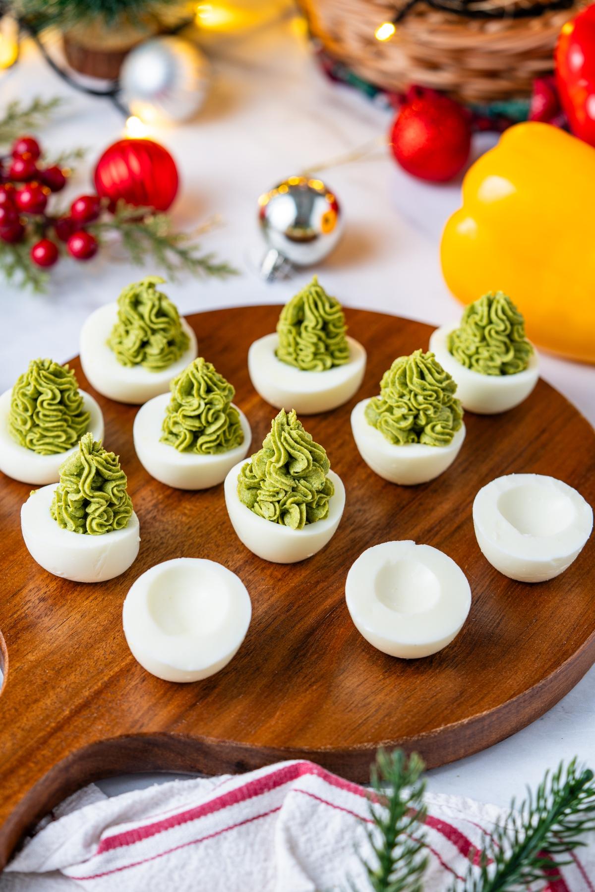 Deviled eggs with green filling piped to look like Christmas trees on a wooden platter, festive decor in background.