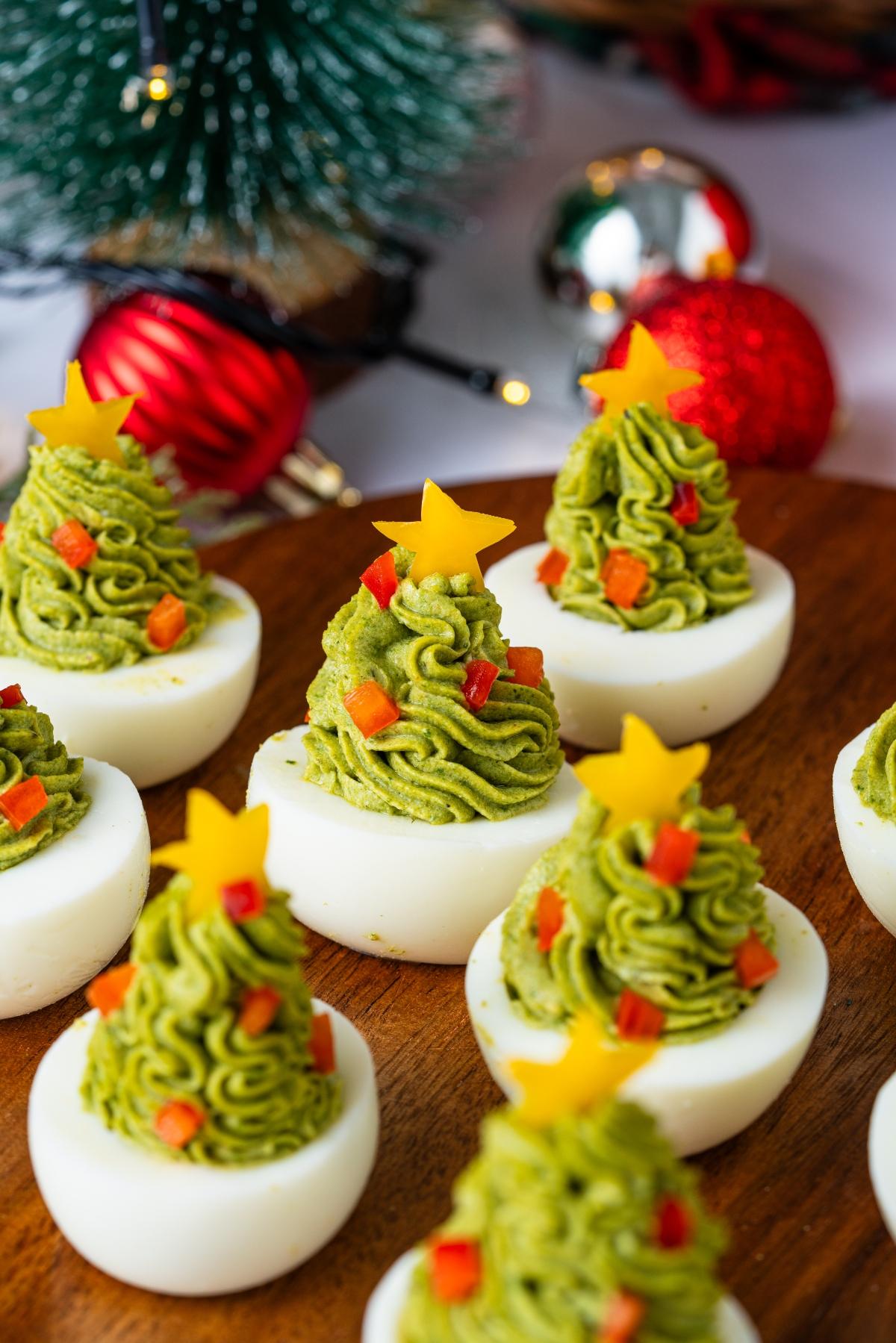 Deviled eggs topped with green filling shaped like Christmas trees, decorated with stars and red bits.