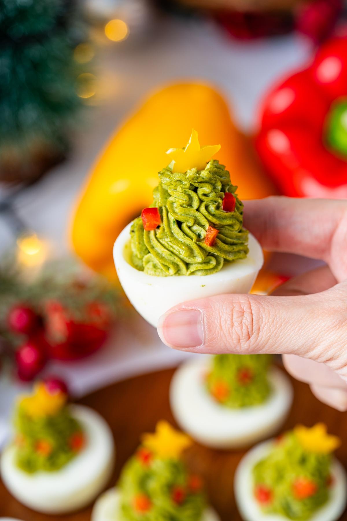 A hand holds a Christmas Tree deviled egg topped with green filling, shaped like a Christmas tree with a yellow star on top.