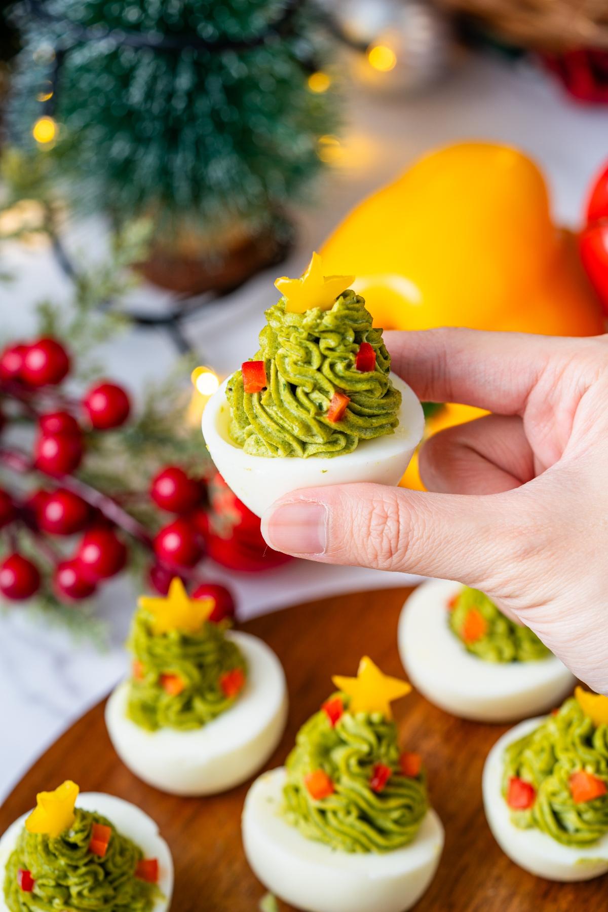 A hand holds a deviled egg topped with green filling shaped like a Christmas tree, garnished with a star.