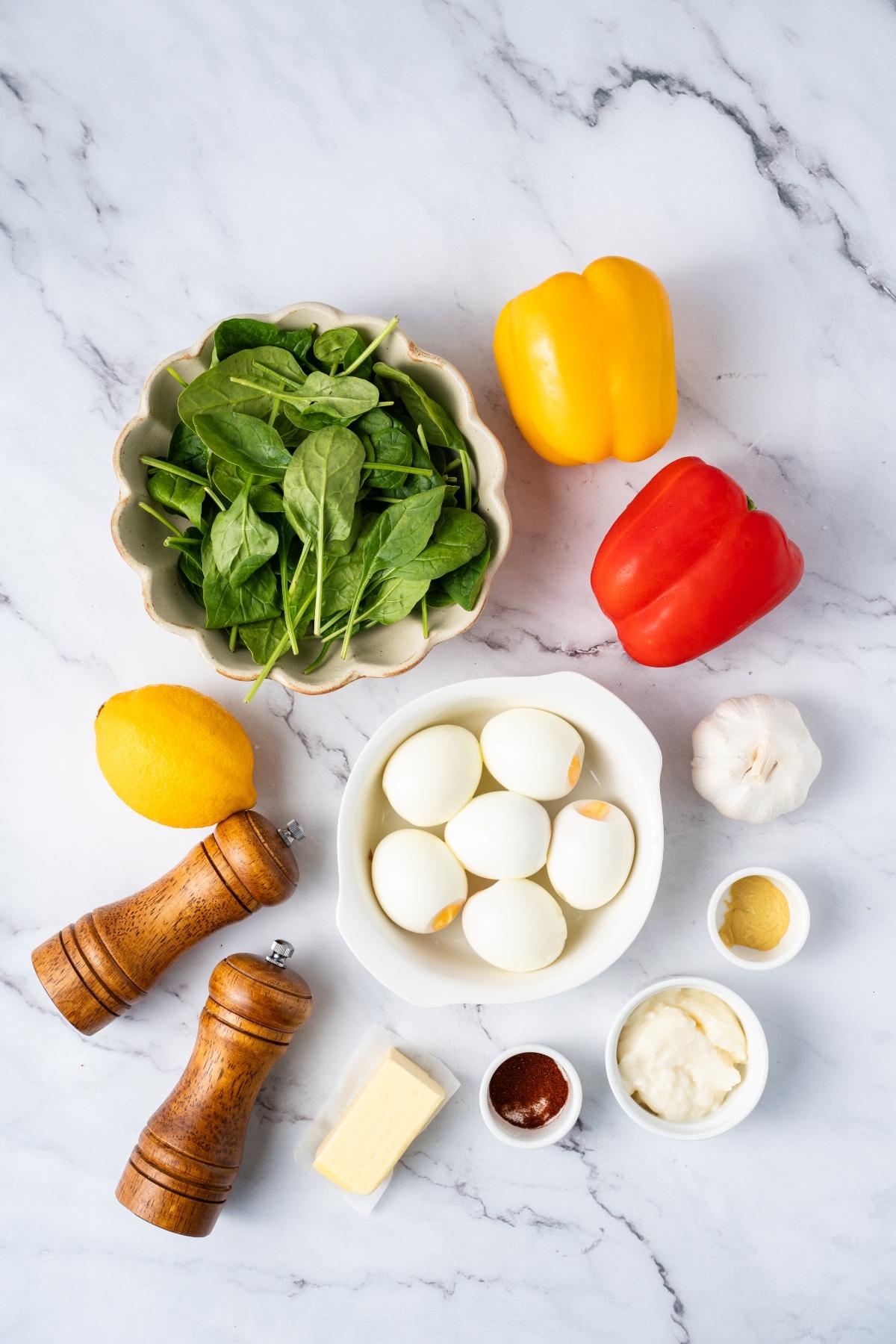 Ingredients on a marble surface: spinach, eggs, bell peppers, lemon, garlic, butter, spices, and condiments.