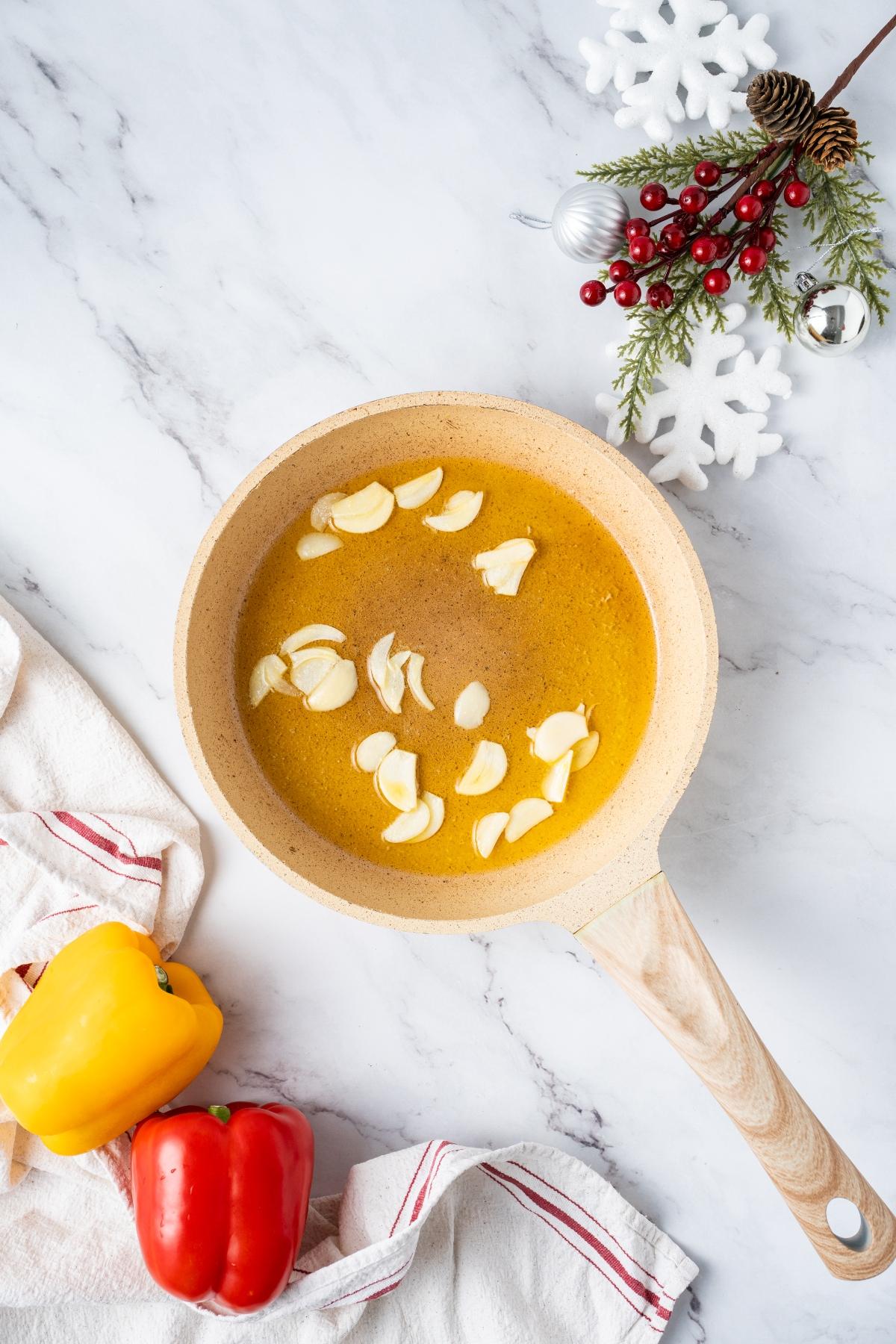 A skillet with oil and sliced garlic, near yellow and red bell peppers, on a marble surface with holiday decor.