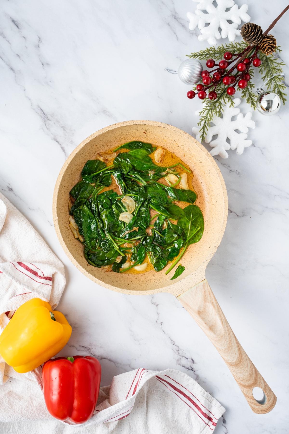 A pan of sautéed spinach and garlic on a marble counter with bell peppers and holiday decorations nearby.