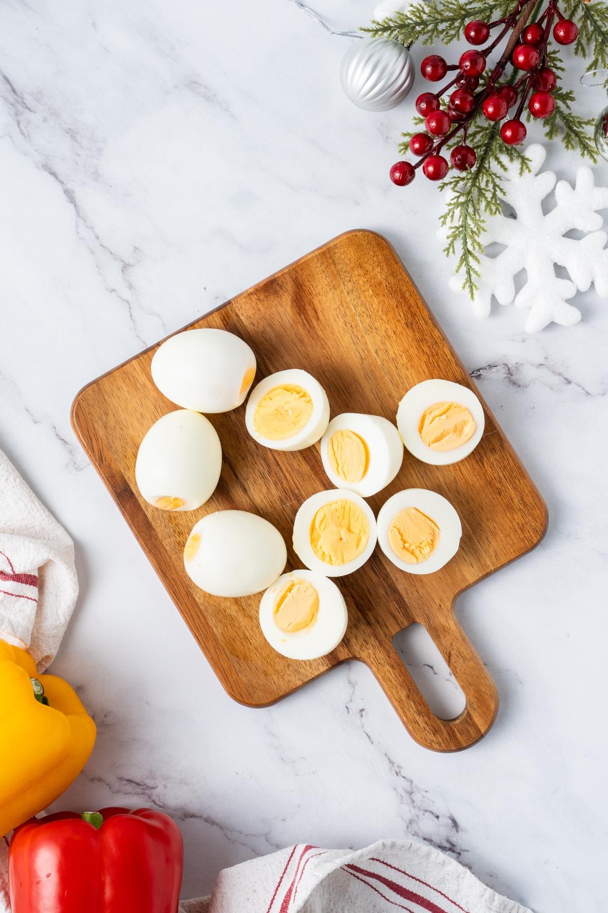 Hard-boiled eggs, whole and halved, on a wooden board with holiday decor on a marble surface.