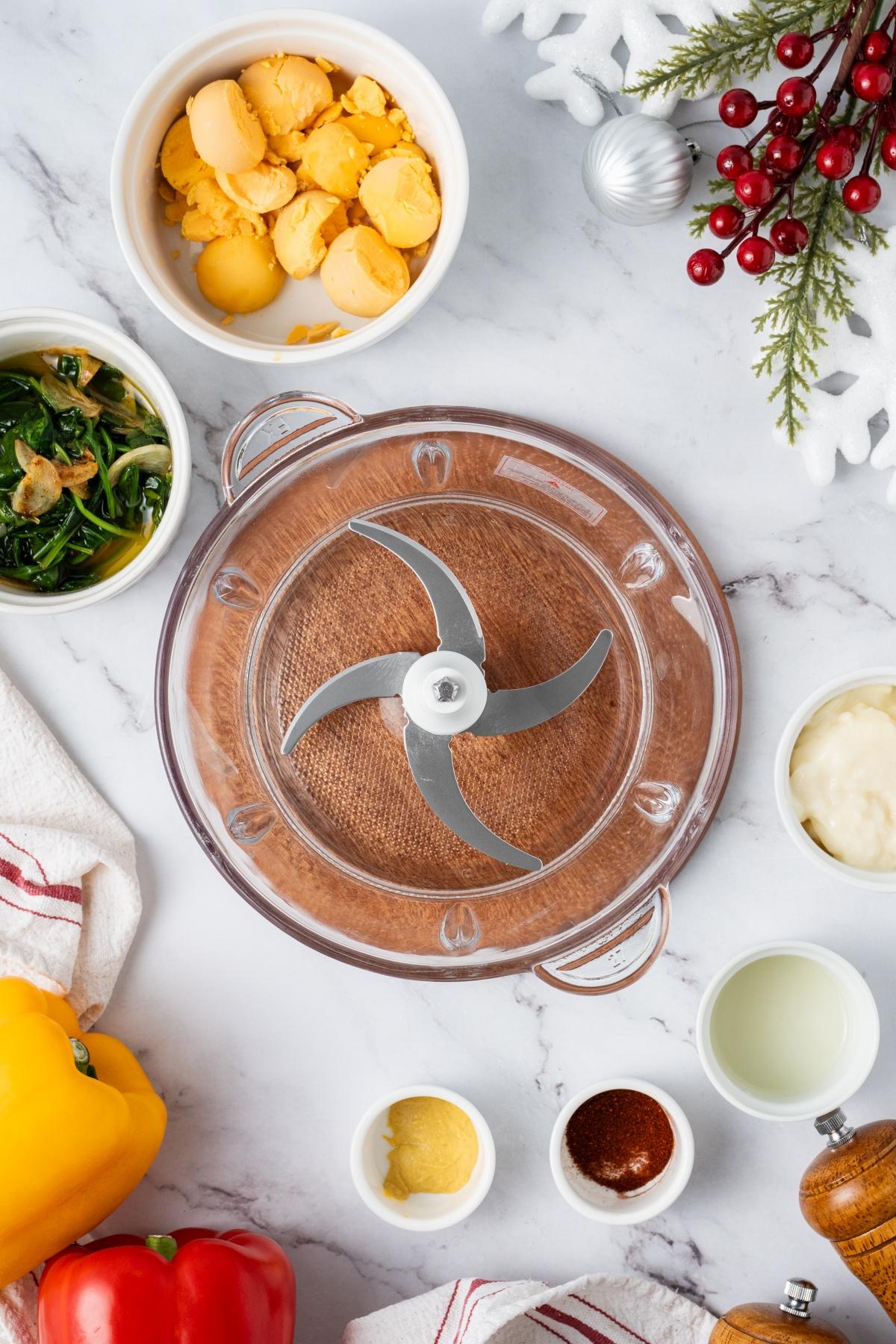 Overhead view of food processor with ingredients like egg yolks, peppers, mustard, and greens on marble surface.