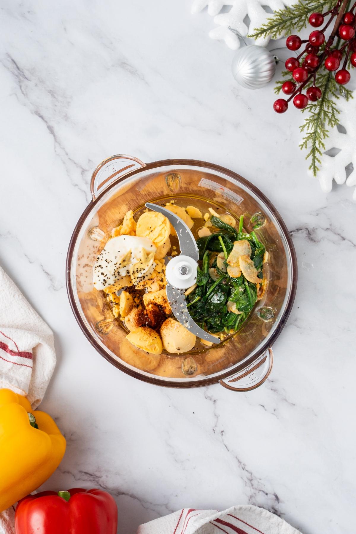 A food processor with chickpeas, spinach, spices, and yogurt on a marble counter with holiday decor nearby.
