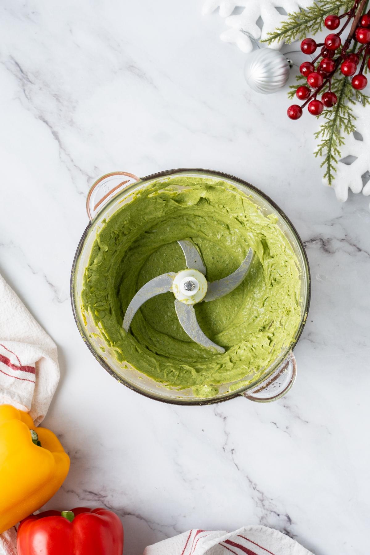 Food processor with creamy green mixture inside, surrounded by holiday decor and bell peppers on a marble surface.