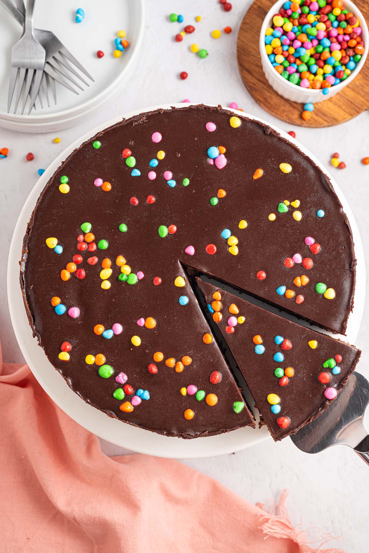 Cosmic Brownie Cheesecake topped with colorful candy sprinkles, one indulgent slice being served, surrounded by scattered candies.